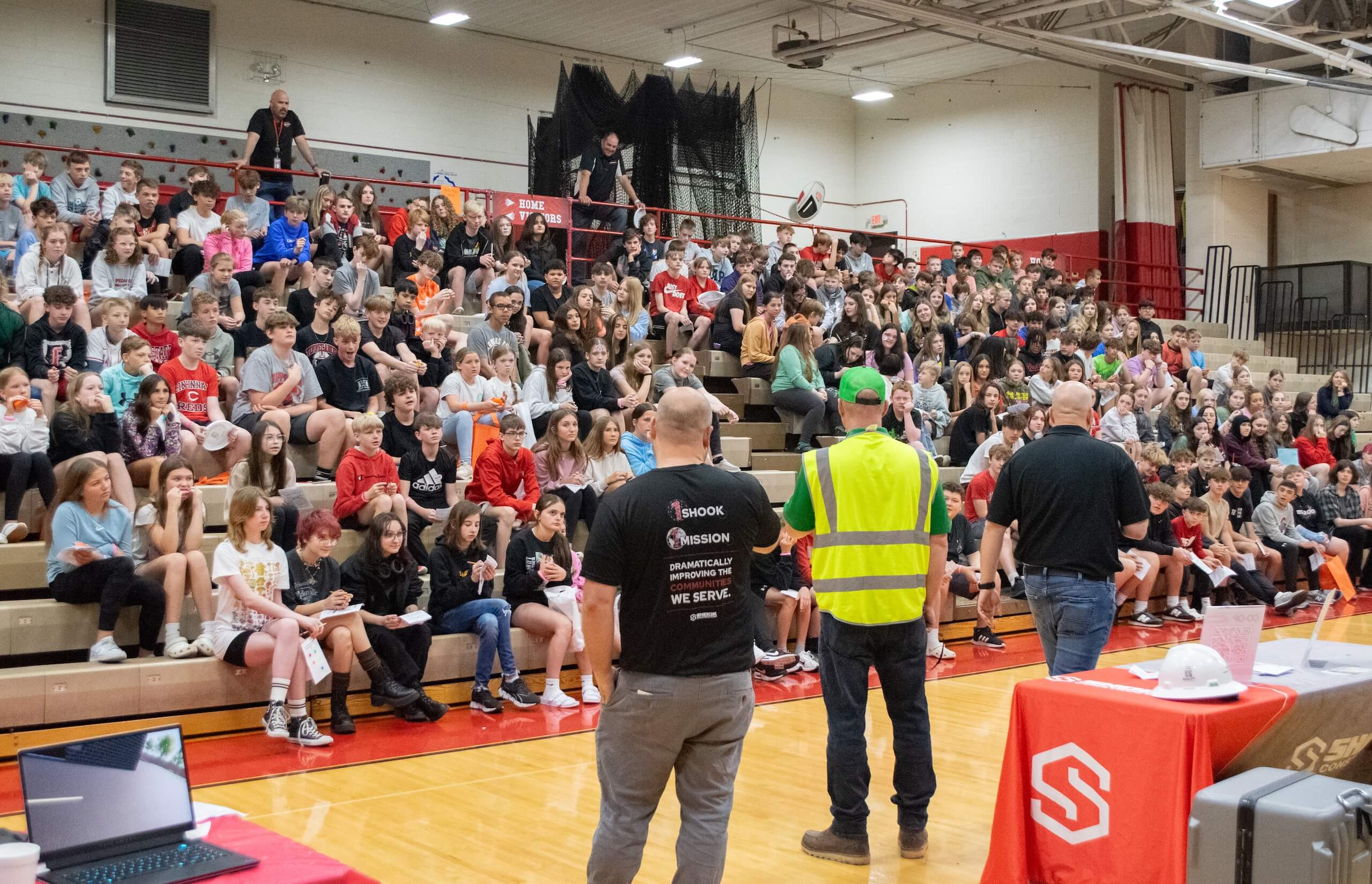Three adults address a large group of students seated on gym bleachers during a school assembly. Some students watch attentively, while others interact with friends. A red table and presentation materials are in the foreground.