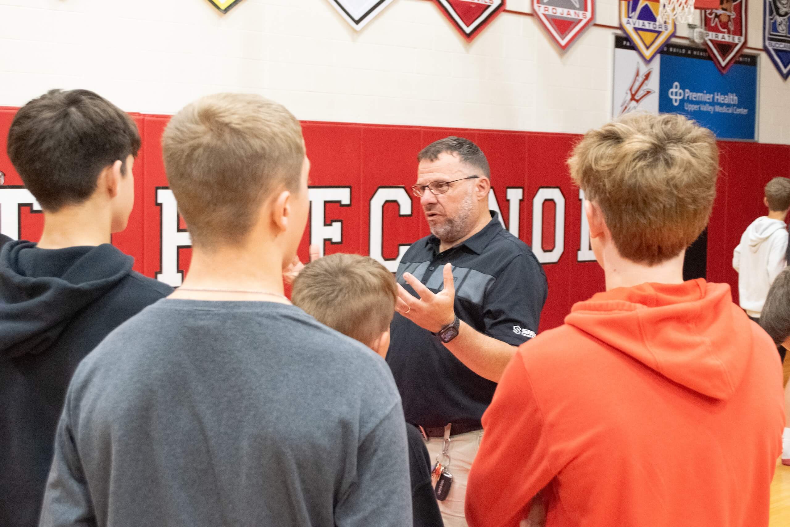 A man speaks to a group of boys in a gymnasium, gesturing with his hands. Banners hang on the wall behind them.