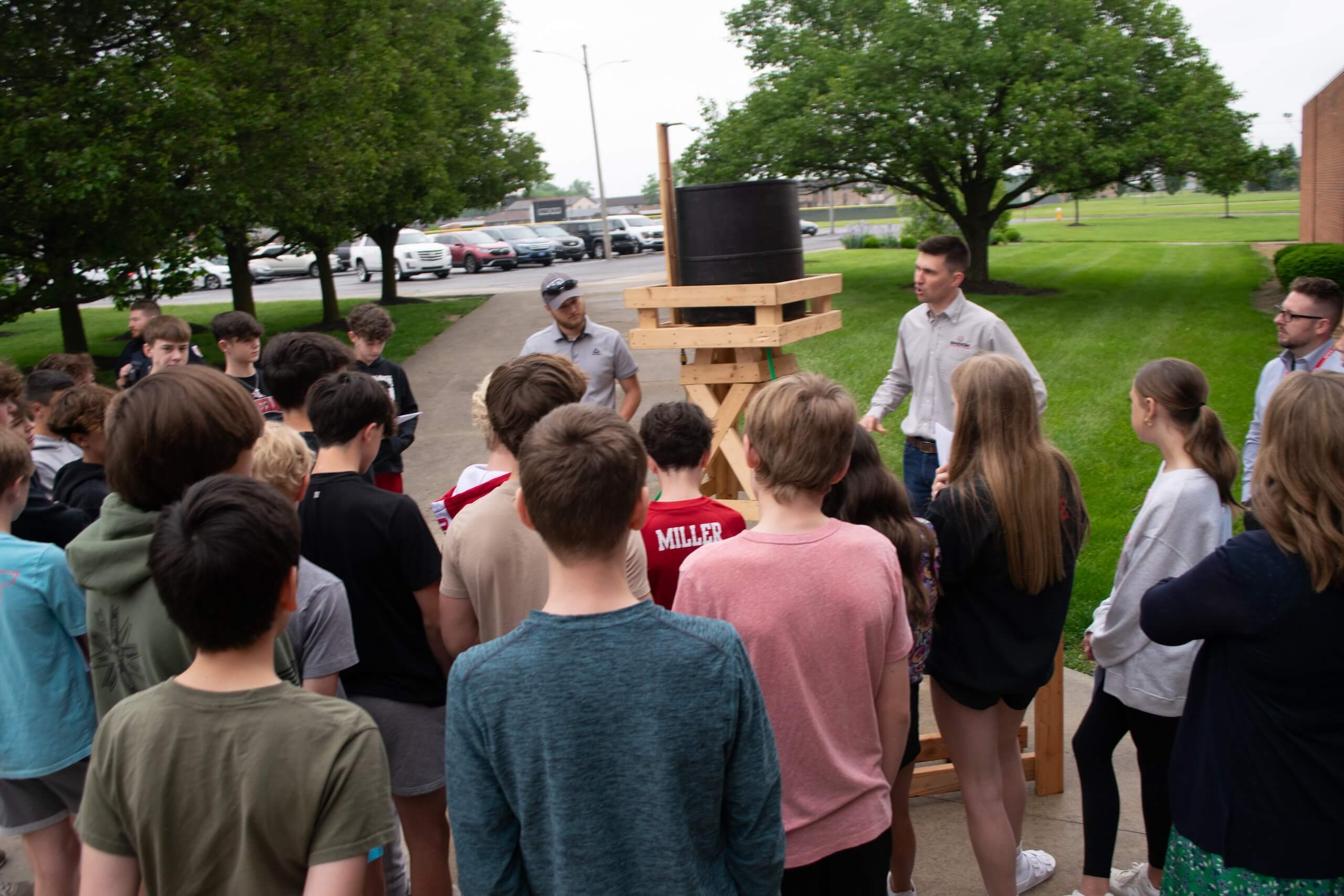 A group of students gathers outdoors around two adults and a wooden contraption with a large black barrel, listening attentively. Trees and parked cars are visible in the background.