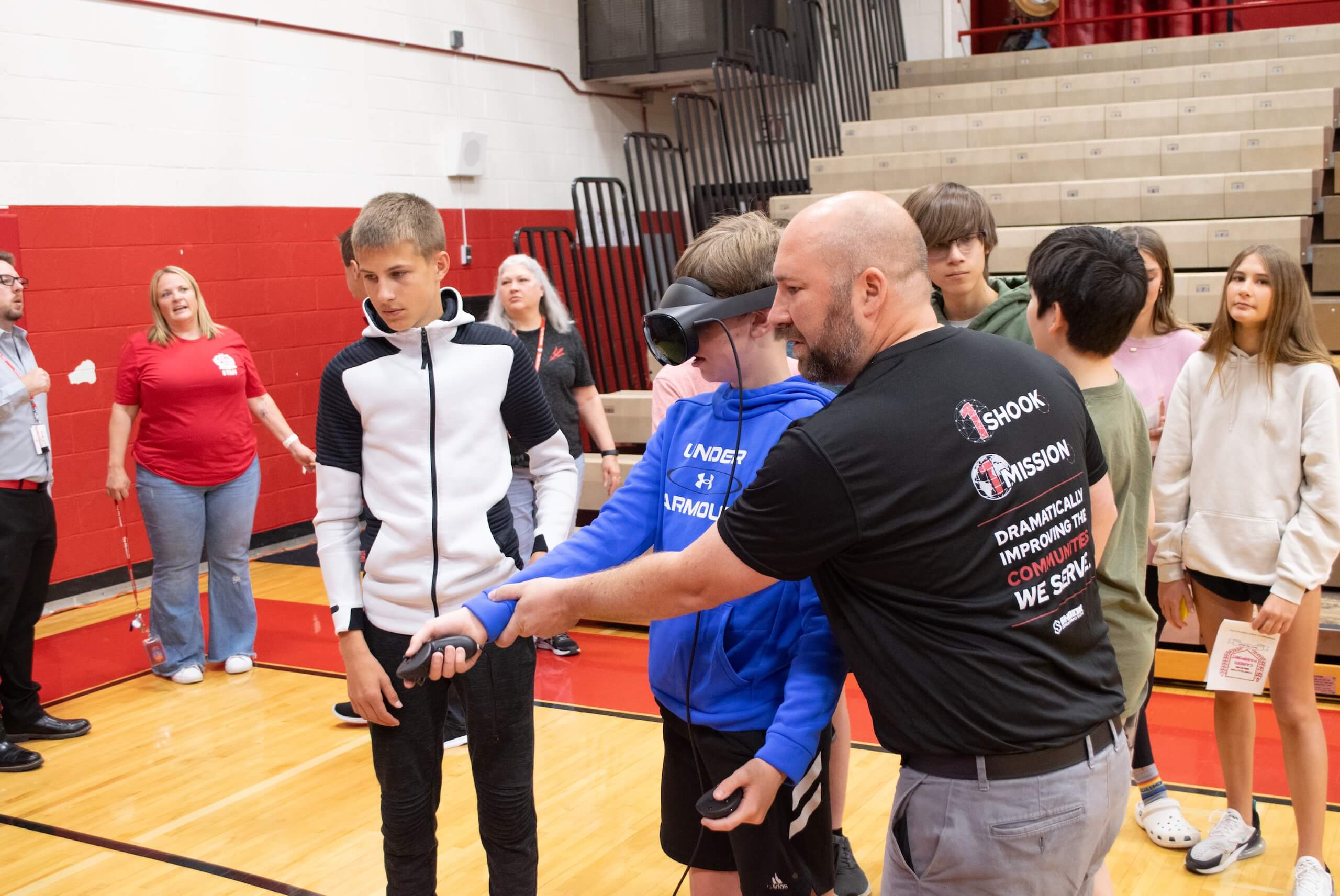 A man helps a student using a VR headset in a gym, surrounded by other students and adults. Some people are observing, and bleachers are visible in the background.