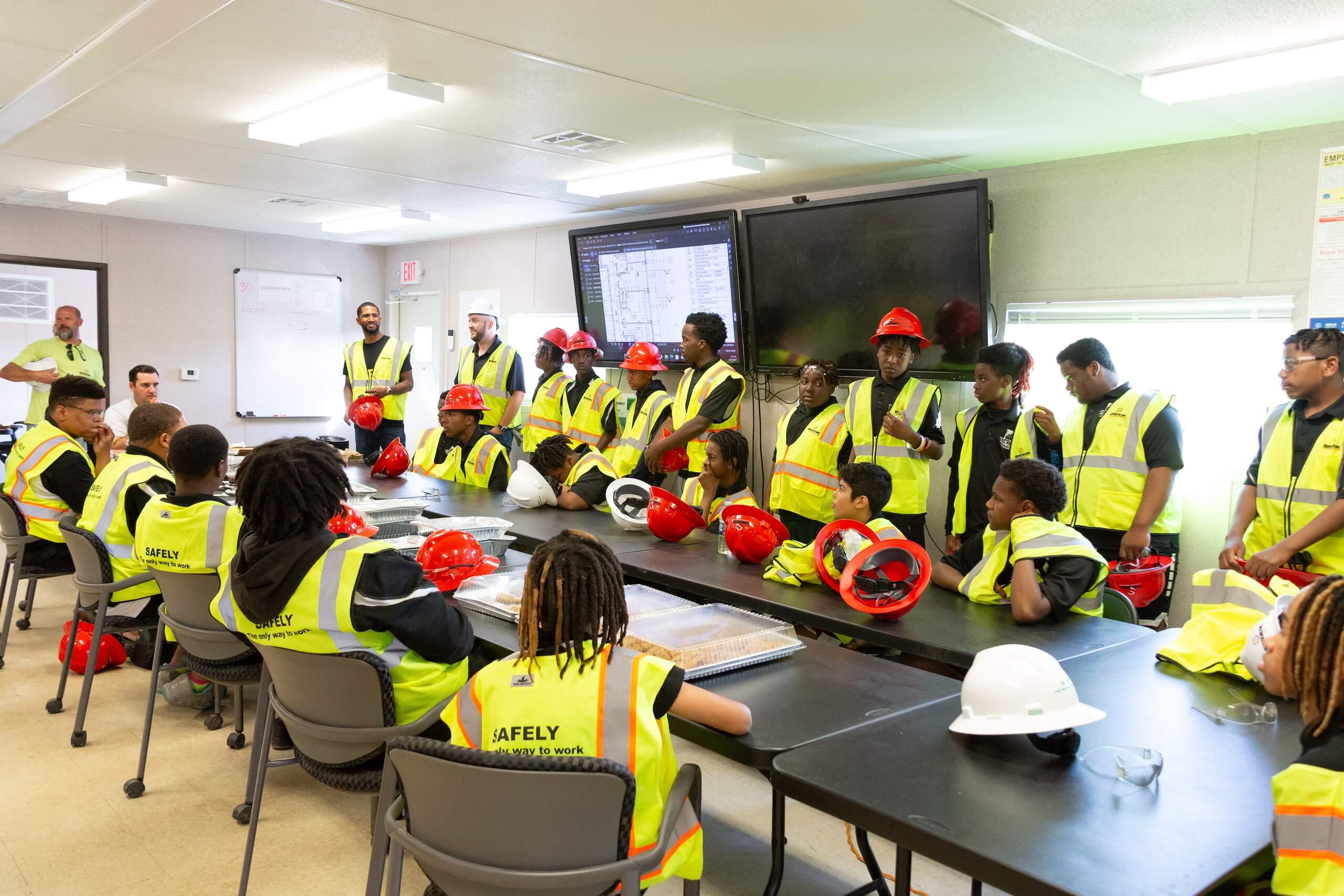 A group of people in safety vests and hard hats attend a meeting in a conference room, sitting and standing around tables with monitors and construction plans visible.