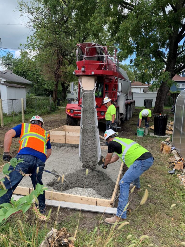 Four construction workers guide wet cement from a truck chute into a wooden frame on the ground, working outdoors near trees and houses.