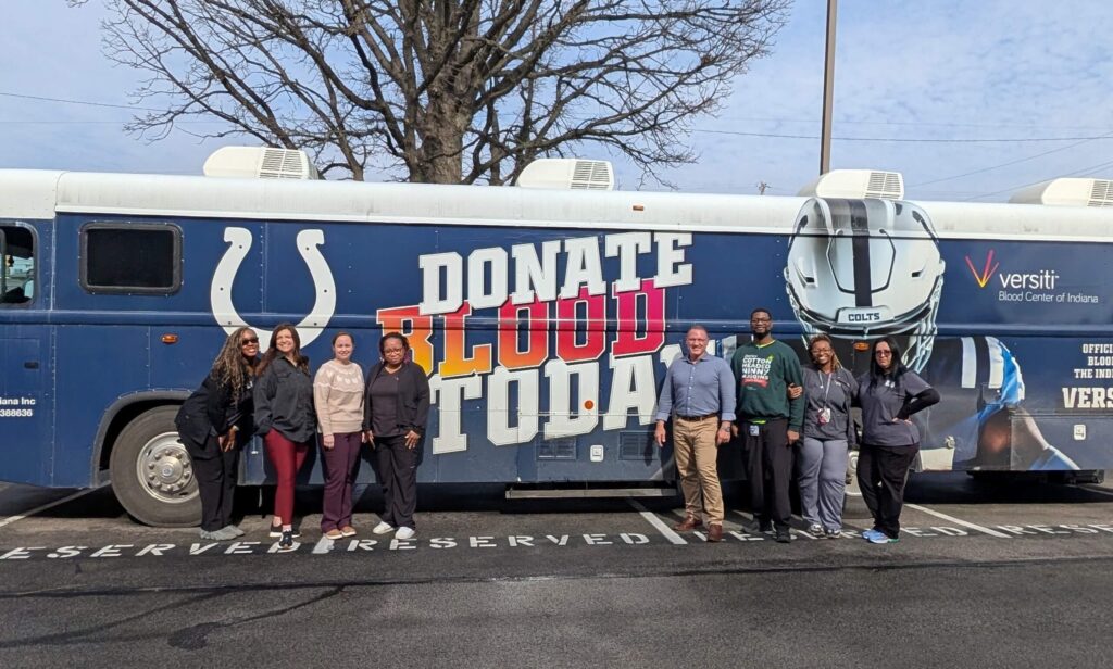 A group of people stand and smile in front of a large bus with a sign that reads Donate Blood Today and displays the Indianapolis Colts logo and branding.