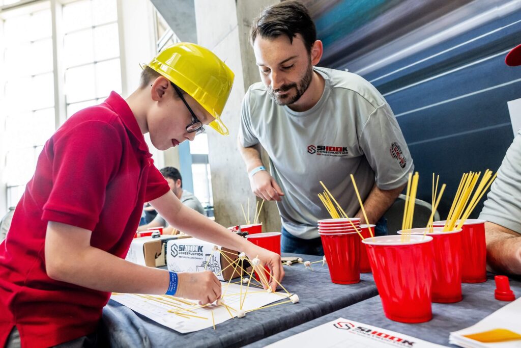 A boy in a yellow hard hat builds a structure with spaghetti and marshmallows while a man watches. Red plastic cups and papers are on the table in a brightly lit room.