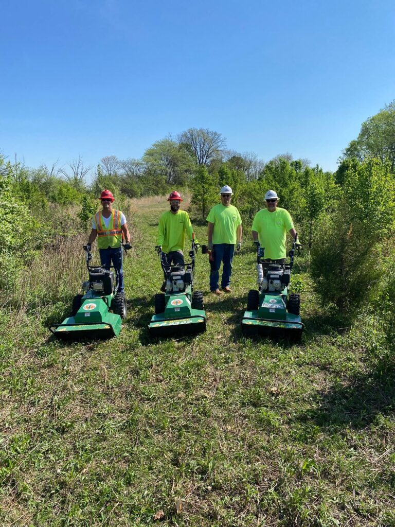 Four people in safety gear and neon shirts stand outdoors on grass, each holding a green walk-behind mower, with trees and bushes in the background under a clear blue sky.