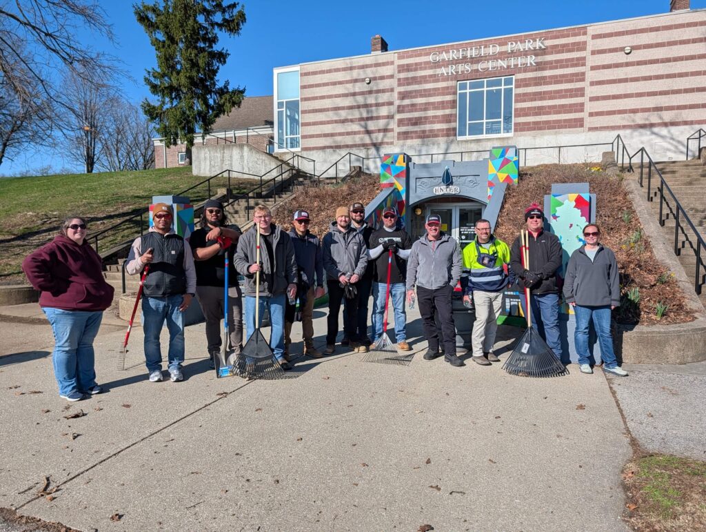 A group of people with rakes and tools pose outside Garfield Park Arts Center on a sunny day, ready for outdoor work or cleanup.