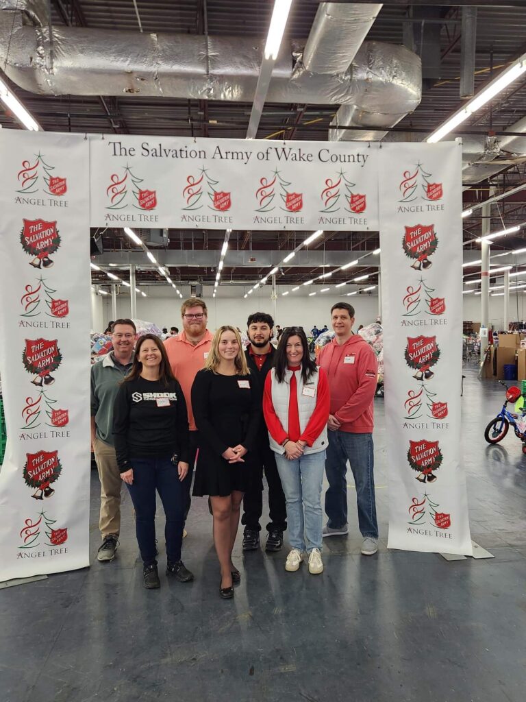 Seven people stand smiling under a banner reading The Salvation Army of Wake County Angel Tree inside a large warehouse with toys visible in the background.