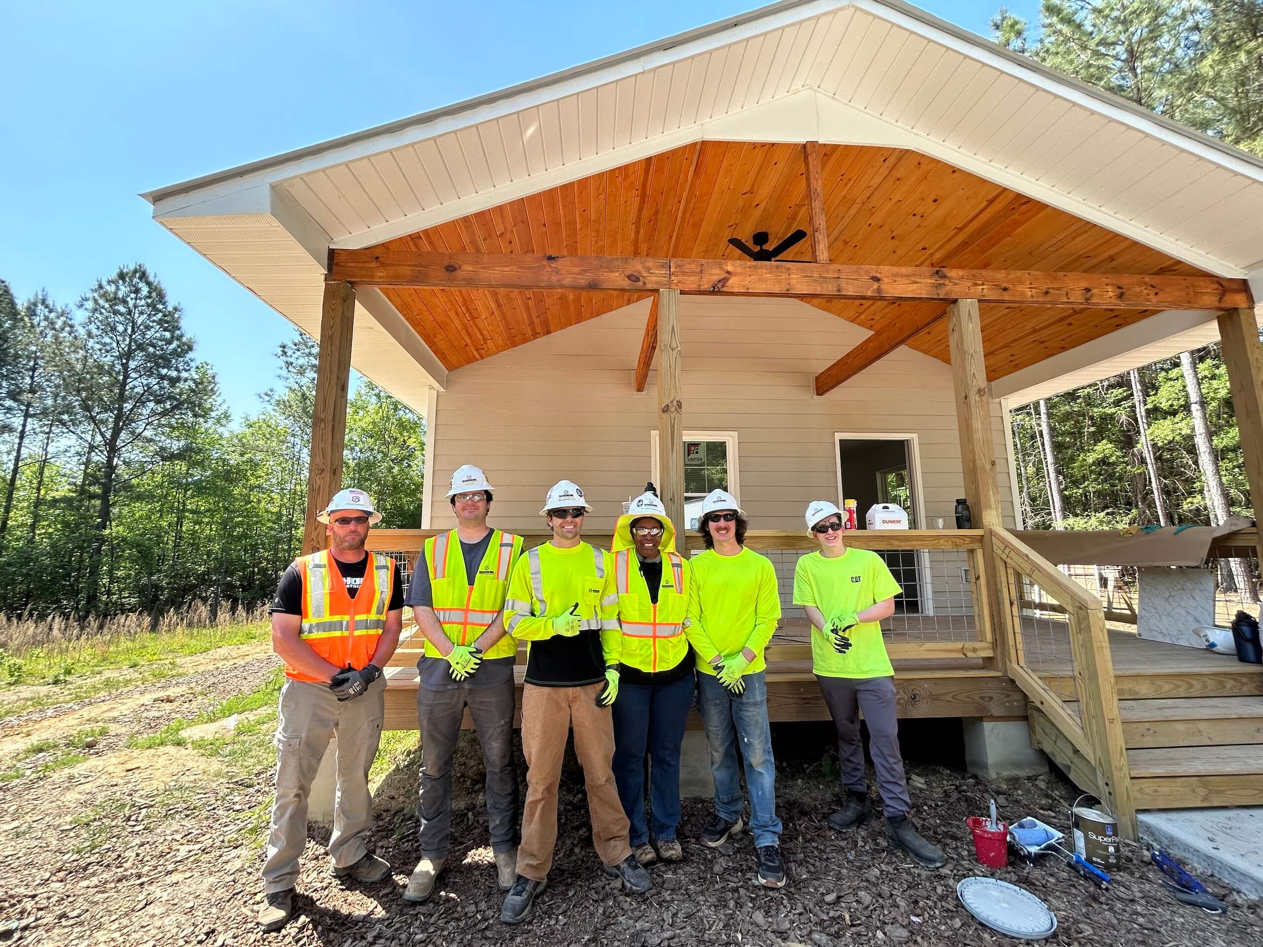 Six people in safety gear and hard hats stand smiling in front of a small house under construction, surrounded by trees and sunlight, with tools and paint supplies visible nearby.