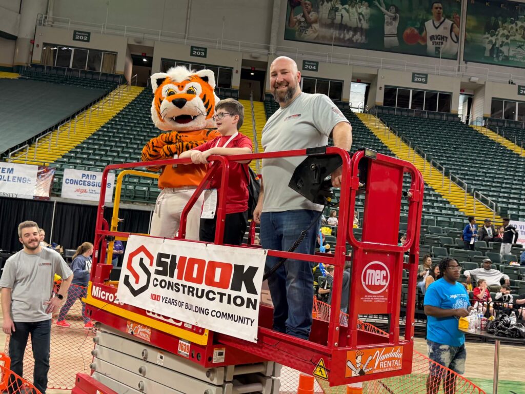 Two men, one in a tiger mascot costume, stand on a red lift with a Shook Construction sign in an indoor arena, surrounded by people and bleachers.