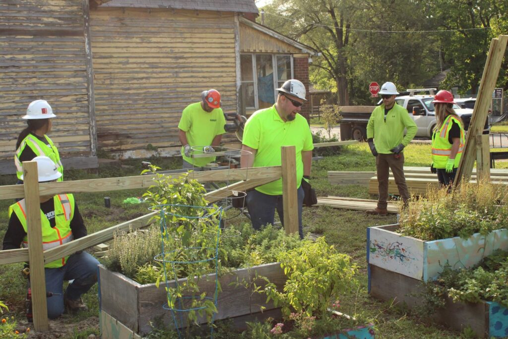 Five construction workers in safety gear and neon shirts work on building a wooden fence near raised garden beds, with an old wooden house and trees in the background.
