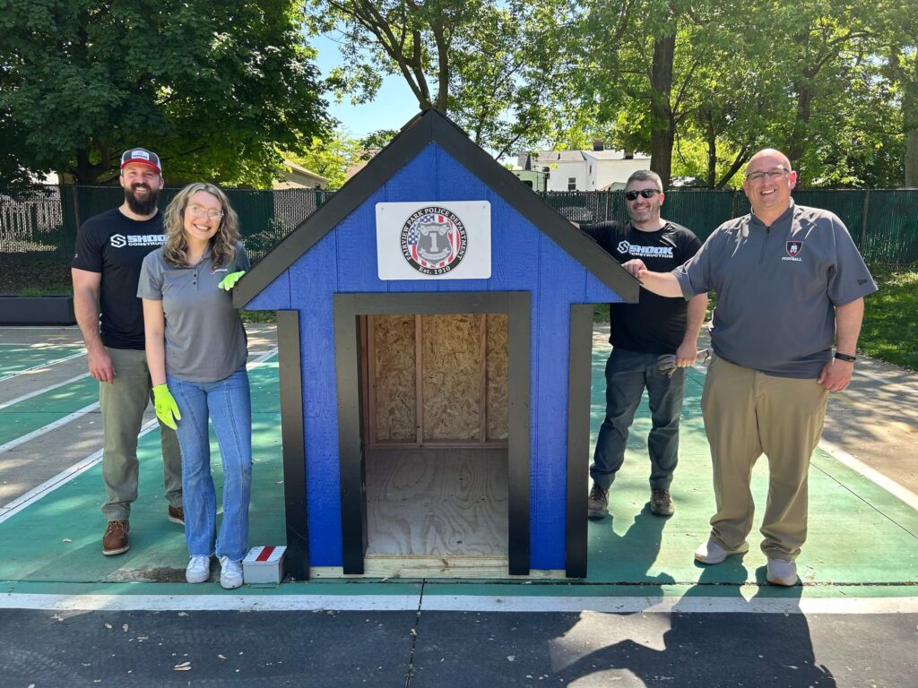 Four smiling adults stand around a blue doghouse on a sunny day. Trees and a fence are in the background. The doghouse features a white sign with a logo above the entrance.