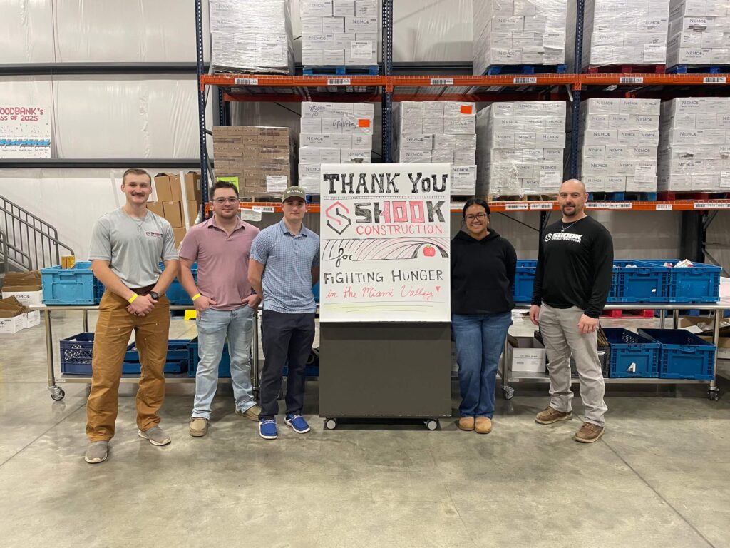 Five people stand in a warehouse with a sign that reads Thank You Shook Construction for fighting hunger in the Miami Valley. Cardboard boxes and blue bins are stacked behind them.