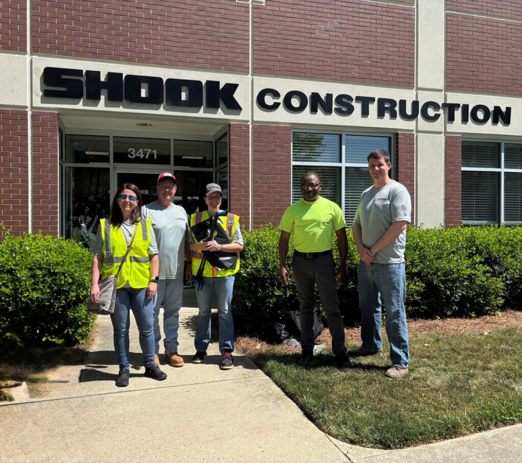 Five people stand smiling in front of a Shook Construction building. Three wear safety vests and one wears a safety shirt. Bushes line the building and the sun is shining.
