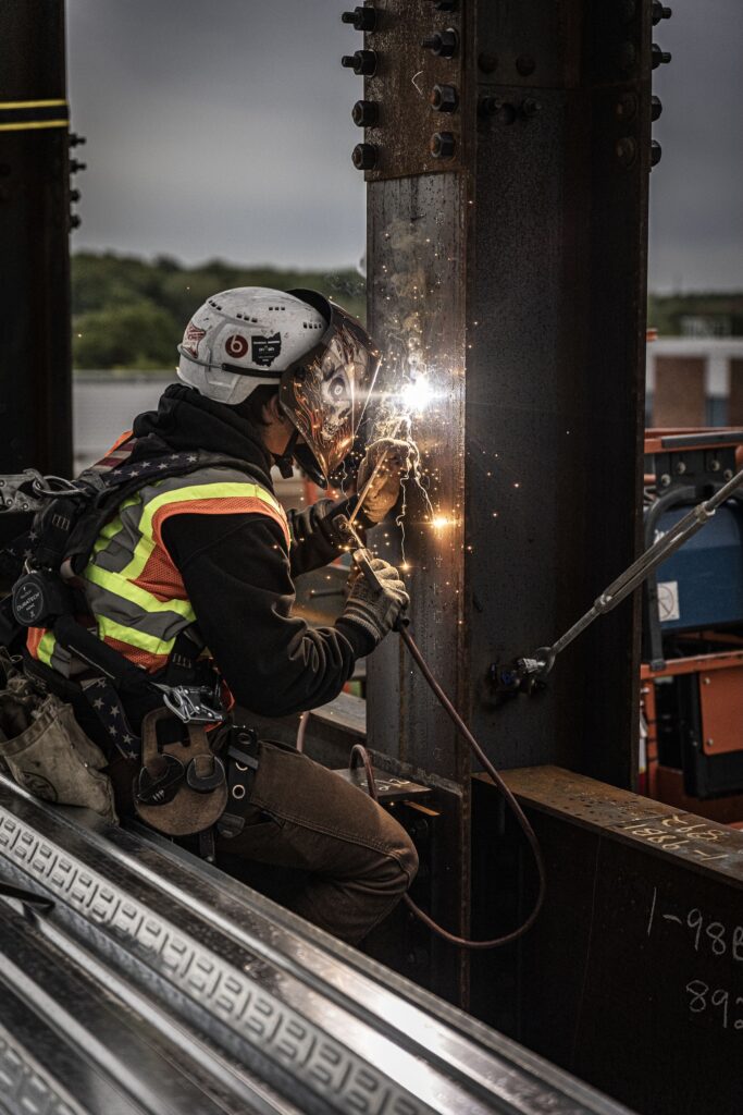 A construction worker wearing a helmet, safety vest, and harness welds a steel beam on a building site, with sparks flying from the welding torch.