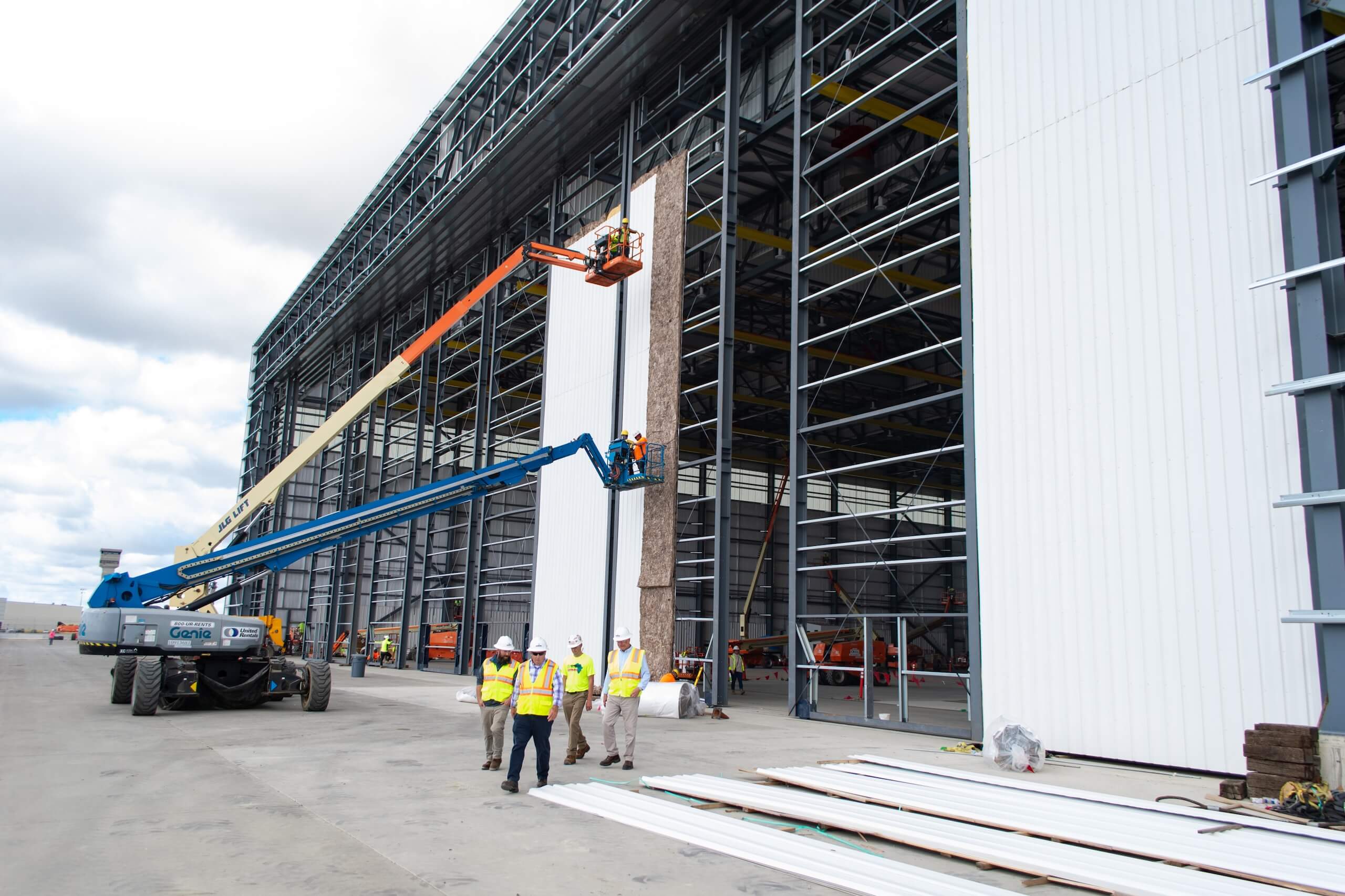 Construction workers in safety vests use lifts to install large white panels on the side of a tall industrial building, with materials and equipment on the ground nearby.