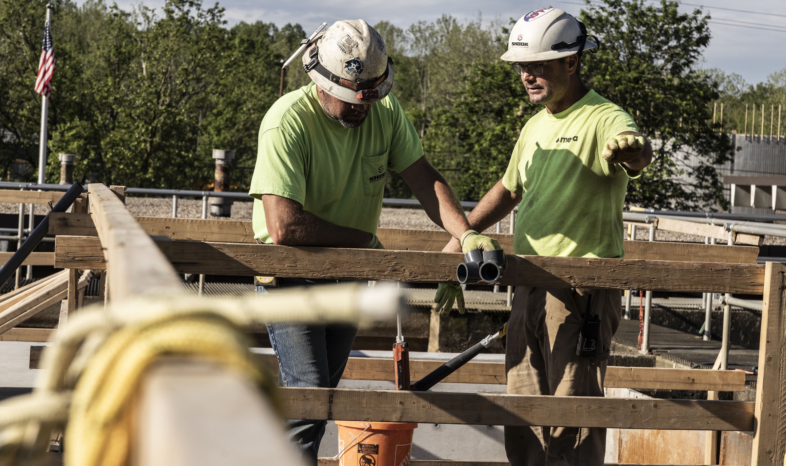 Two construction workers in safety gear discuss plans while building a wooden structure outdoors. One points ahead, and the other looks down at the beams. Trees and an American flag are visible in the background.