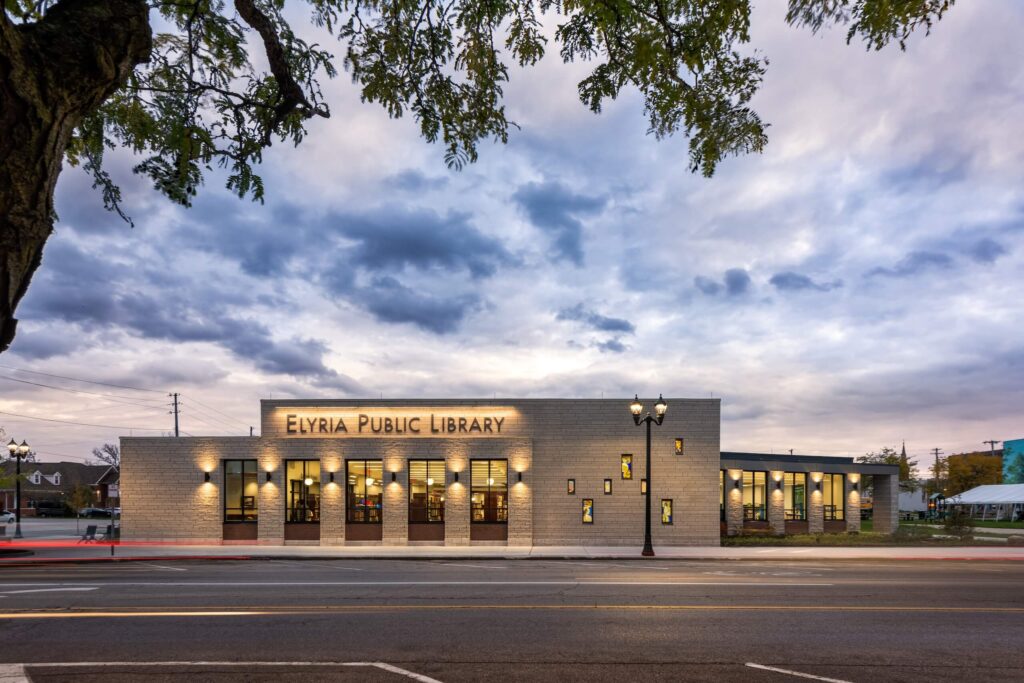 Elyria Public Library building at dusk, illuminated by exterior lights, with a dramatic cloudy sky in the background and a tree branch partially framing the scene.