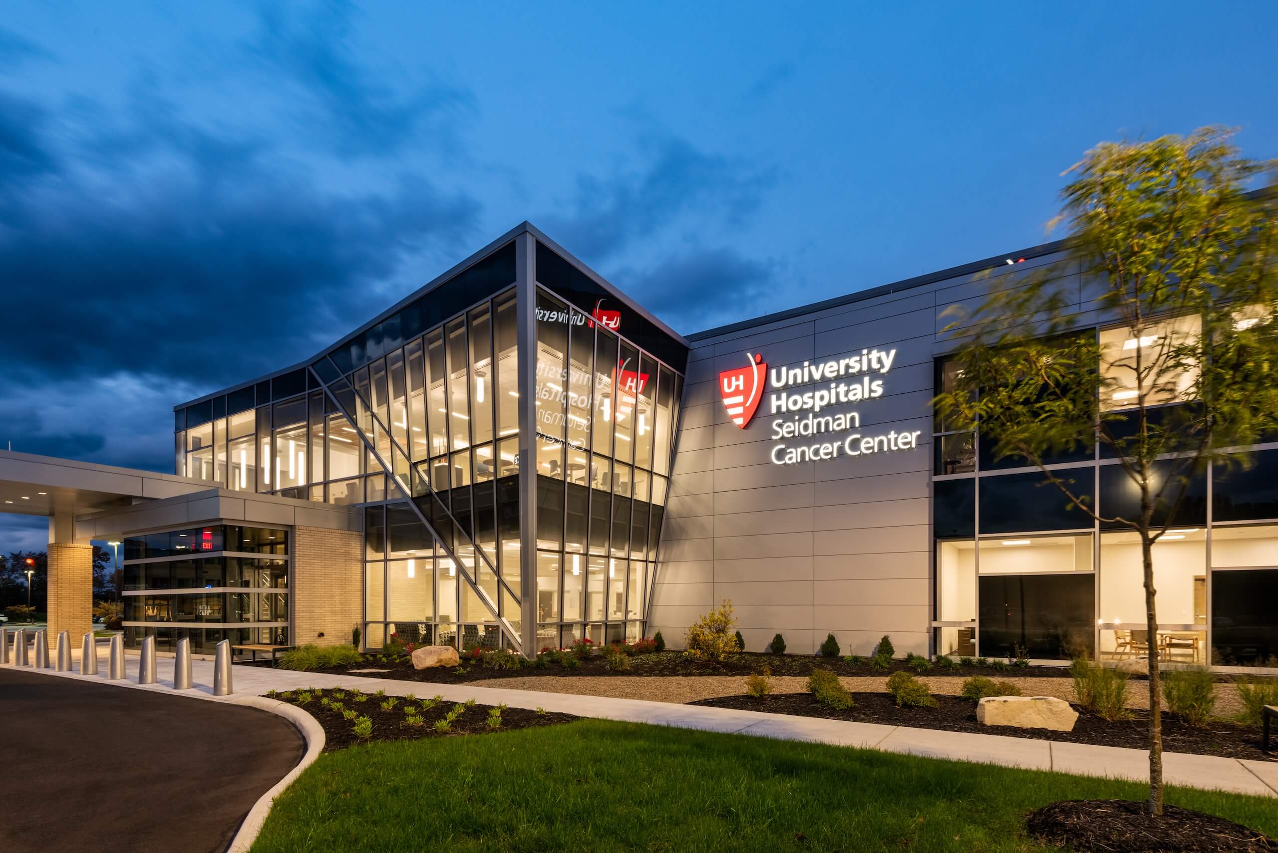 Modern hospital building at dusk with University Hospitals Seidman Cancer Center sign illuminated on the exterior; landscaped grounds and cloudy evening sky in the background.