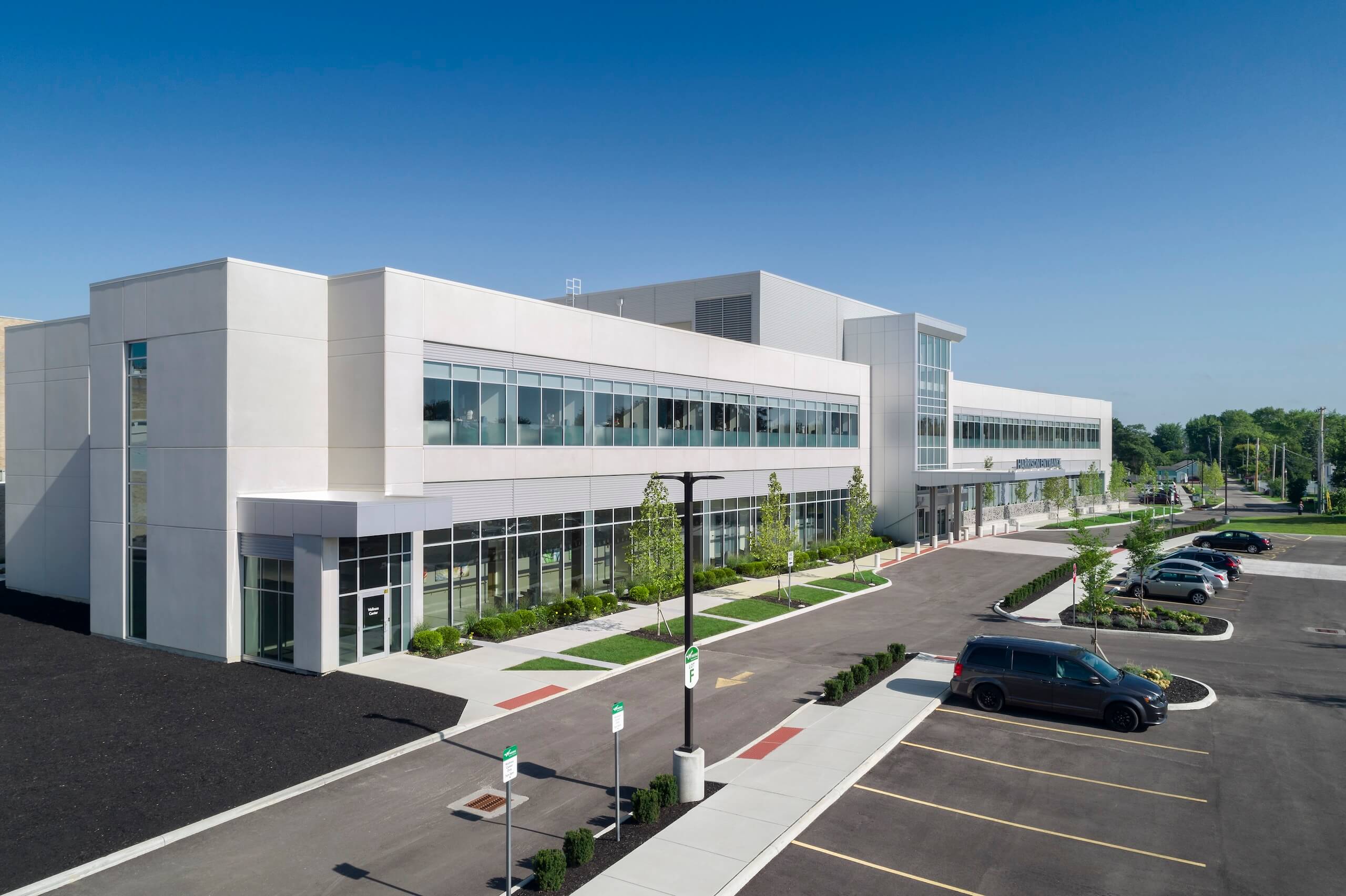 Modern white building with large windows, surrounded by a parking lot with several cars, green landscaping, and a clear blue sky in the background.