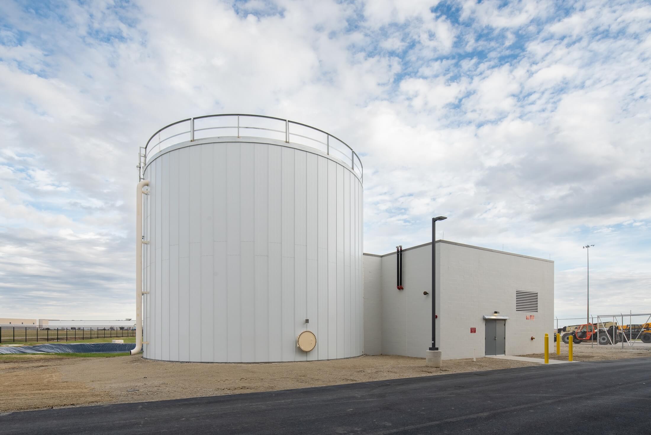 A large white cylindrical storage tank stands next to a small square utility building under a cloudy sky, with an asphalt road and fenced area in the background.