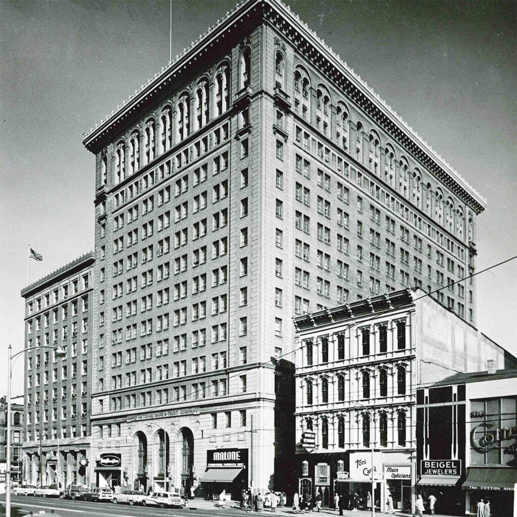 A tall, historic building with arched windows at the base, surrounded by smaller shops and businesses on a busy city street. Pedestrians and cars are visible in front of the building.