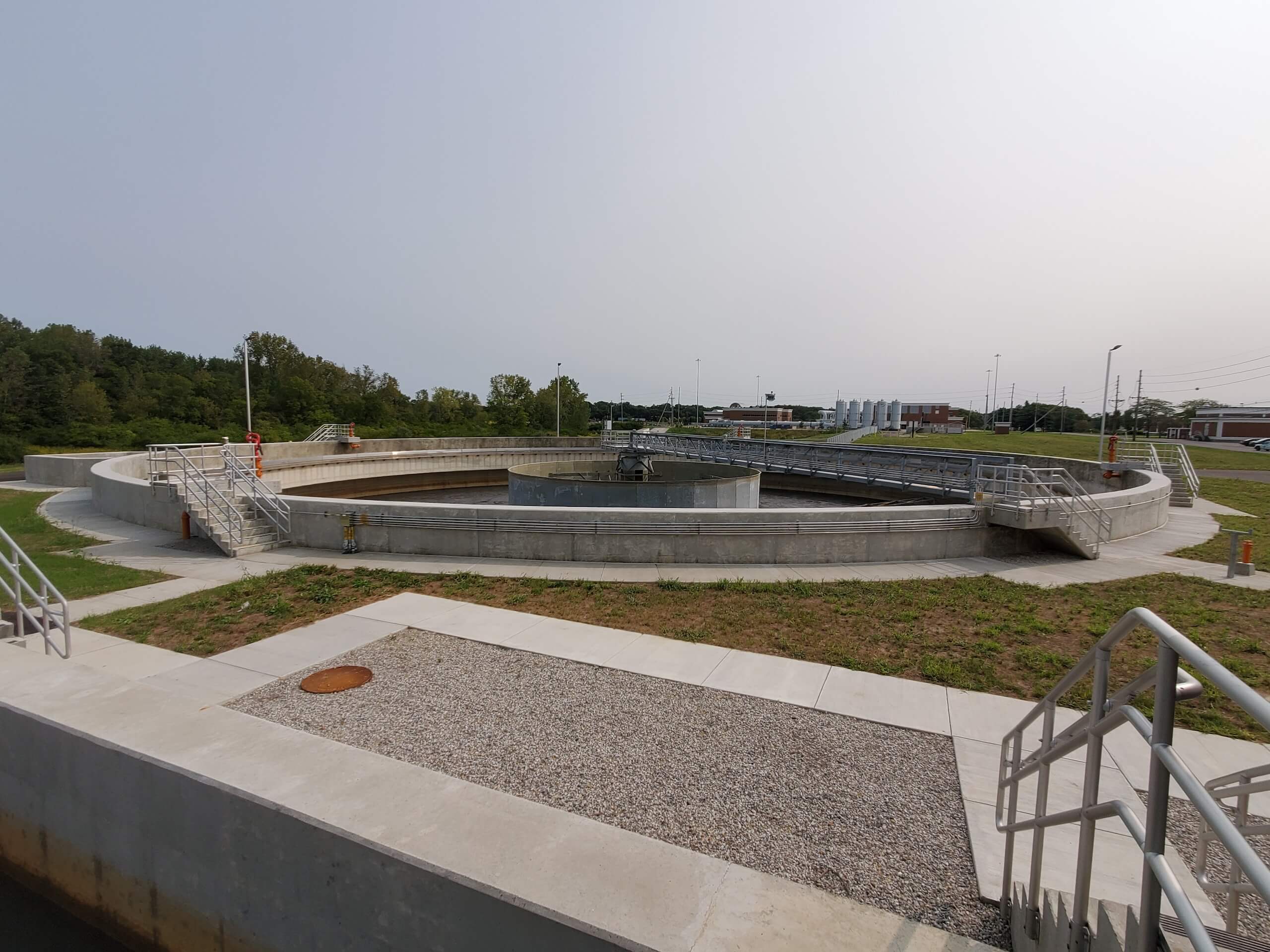 A large, circular water treatment structure with metal railings, surrounded by greenery and buildings in the background, under a clear sky.