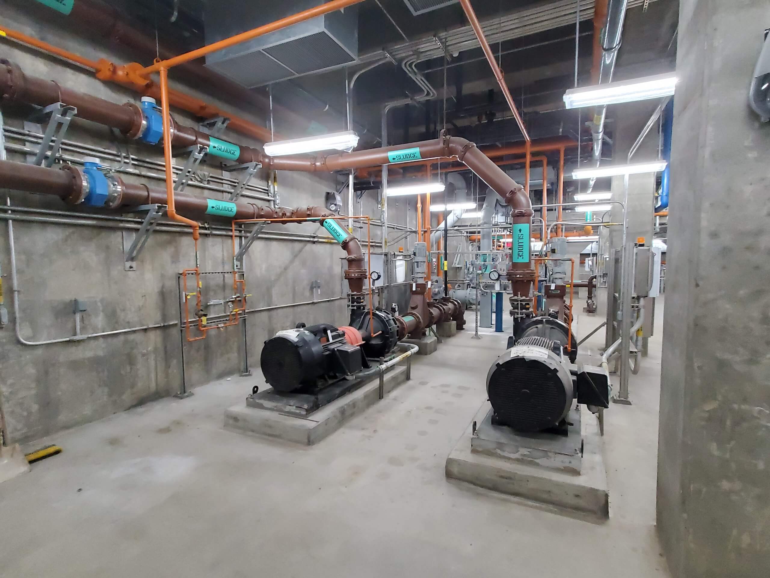 Industrial room with large pumps, pipes, control panels, and exposed wiring on concrete walls. Orange and blue pipes run along the ceiling and walls, with labels attached. The space appears clean and well-lit.