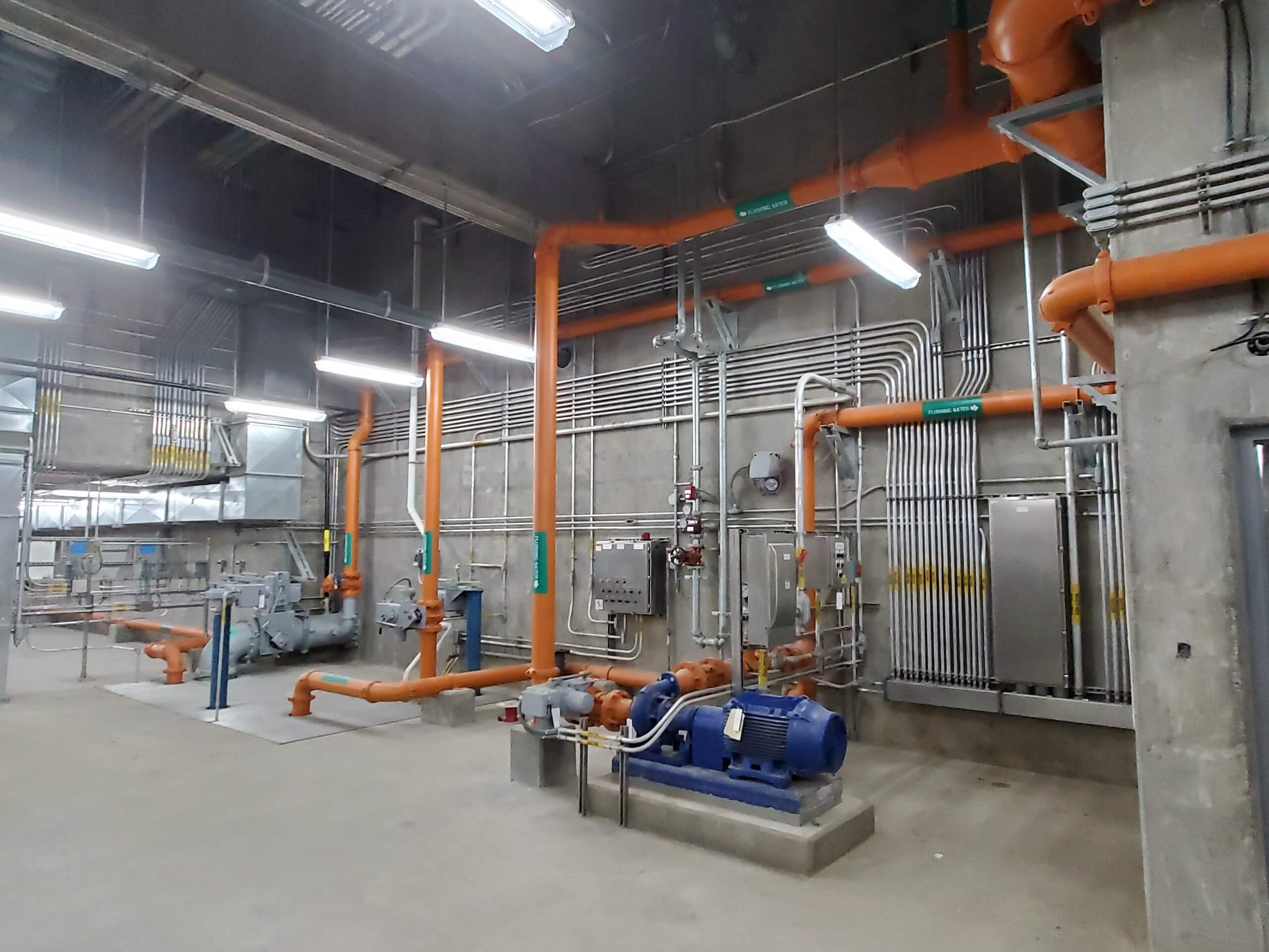 Industrial room with concrete walls, orange and silver pipes, electrical panels, conduit, and machinery including pumps and motors. Overhead lights illuminate the clean, organized workspace.