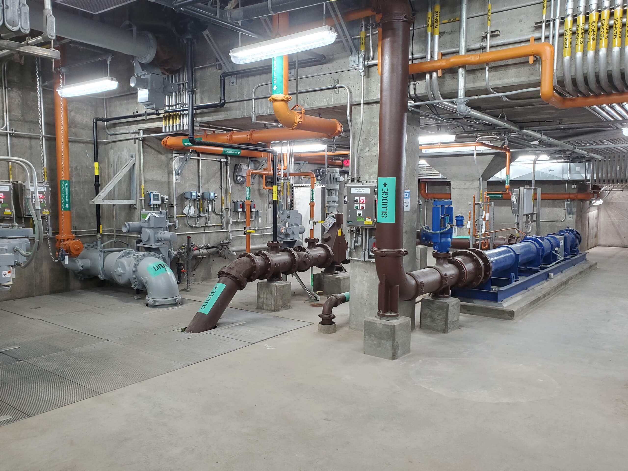 Industrial room with large pipes, pumps, and machinery; labeled pipes run along the floor and ceiling, surrounded by various meters, controls, and exposed wiring on concrete walls. Bright overhead lights illuminate the space.