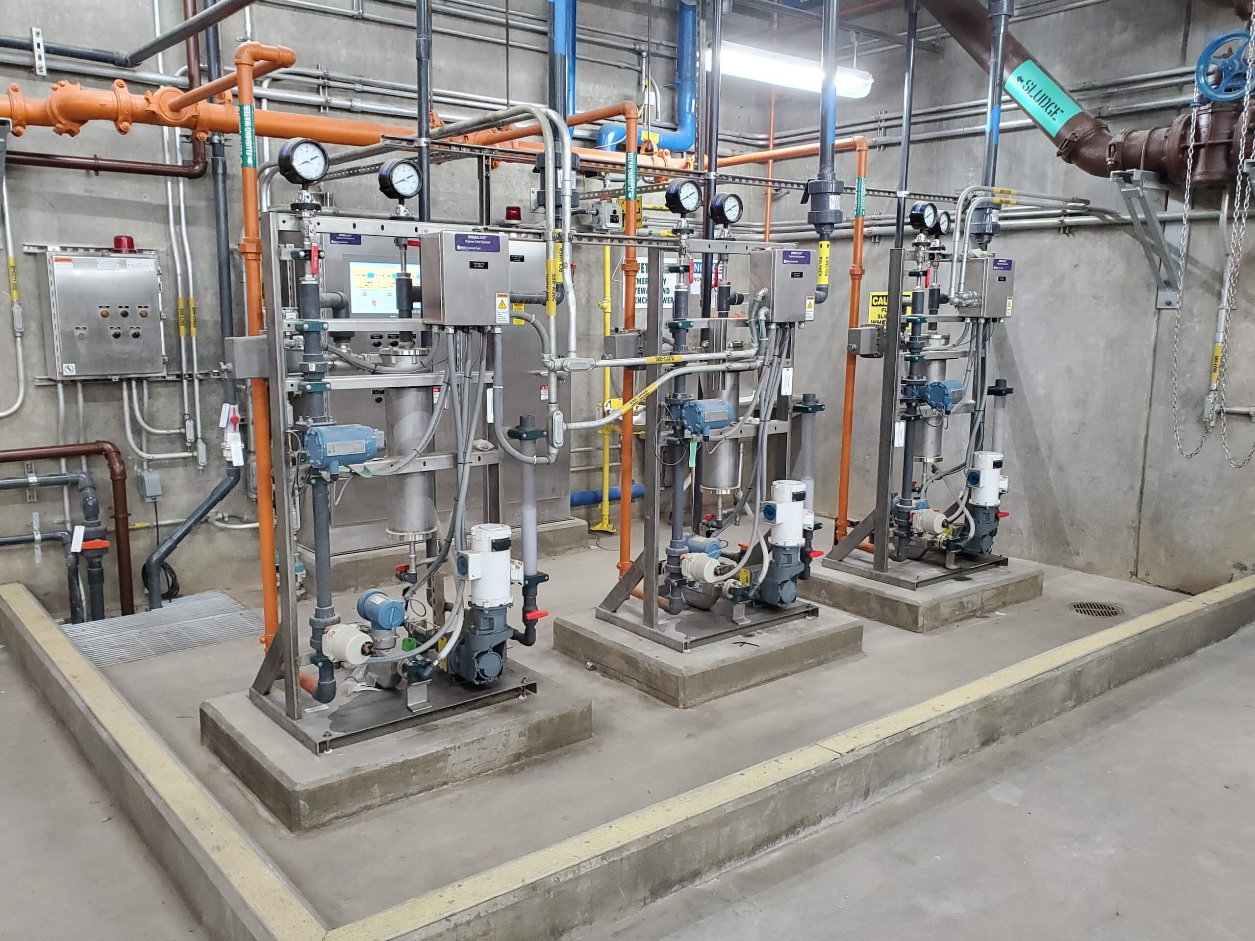 Three industrial pump stations with gauges, pipes, and control panels are installed on concrete platforms in a clean, well-lit utility room with exposed pipes, valves, and electrical boxes on the walls.