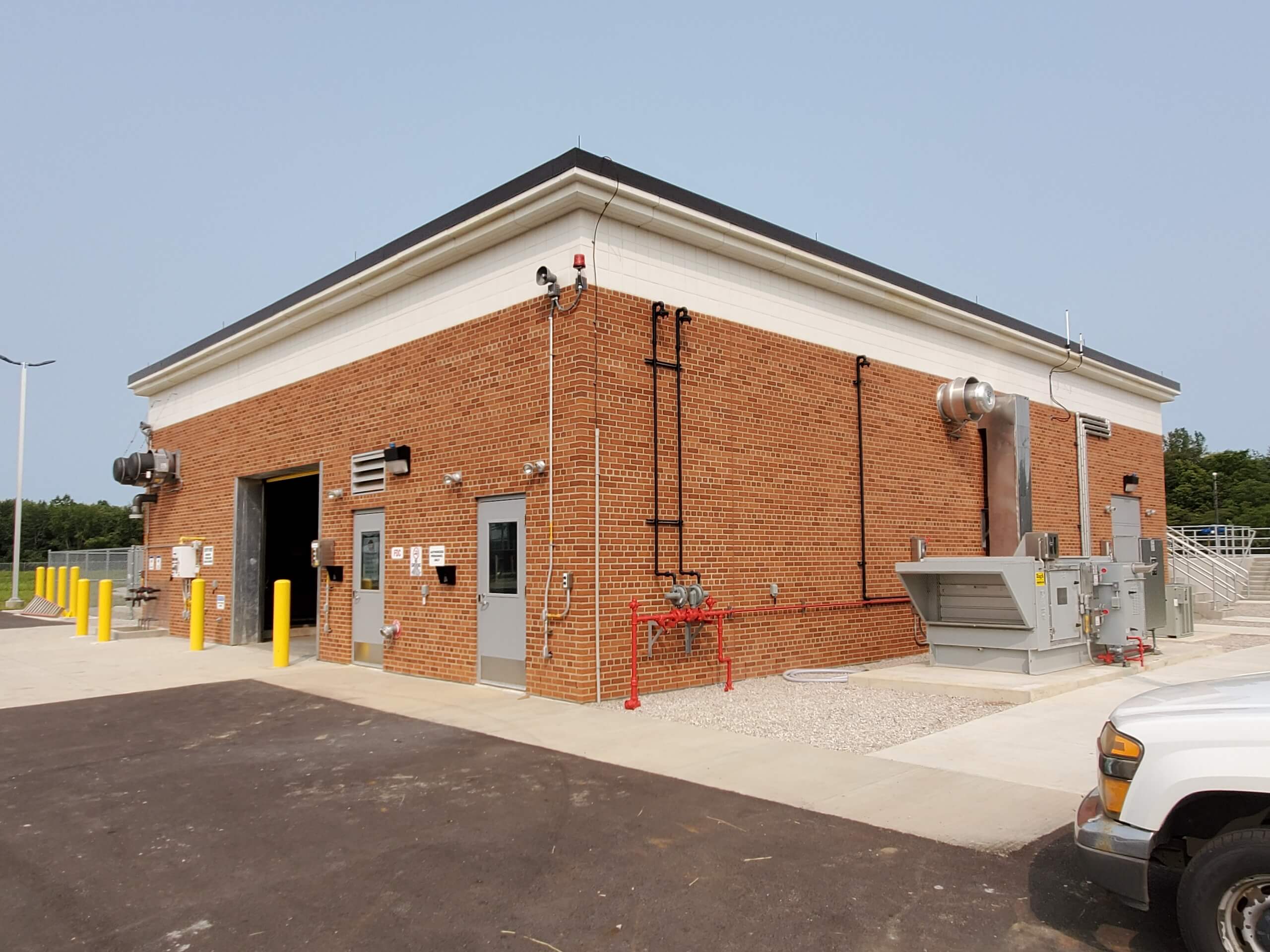 A brick utility building with metal doors, vents, and pipes on the exterior, surrounded by paved roads, yellow bollards, and some vehicles. The sky is clear and the area appears industrial.