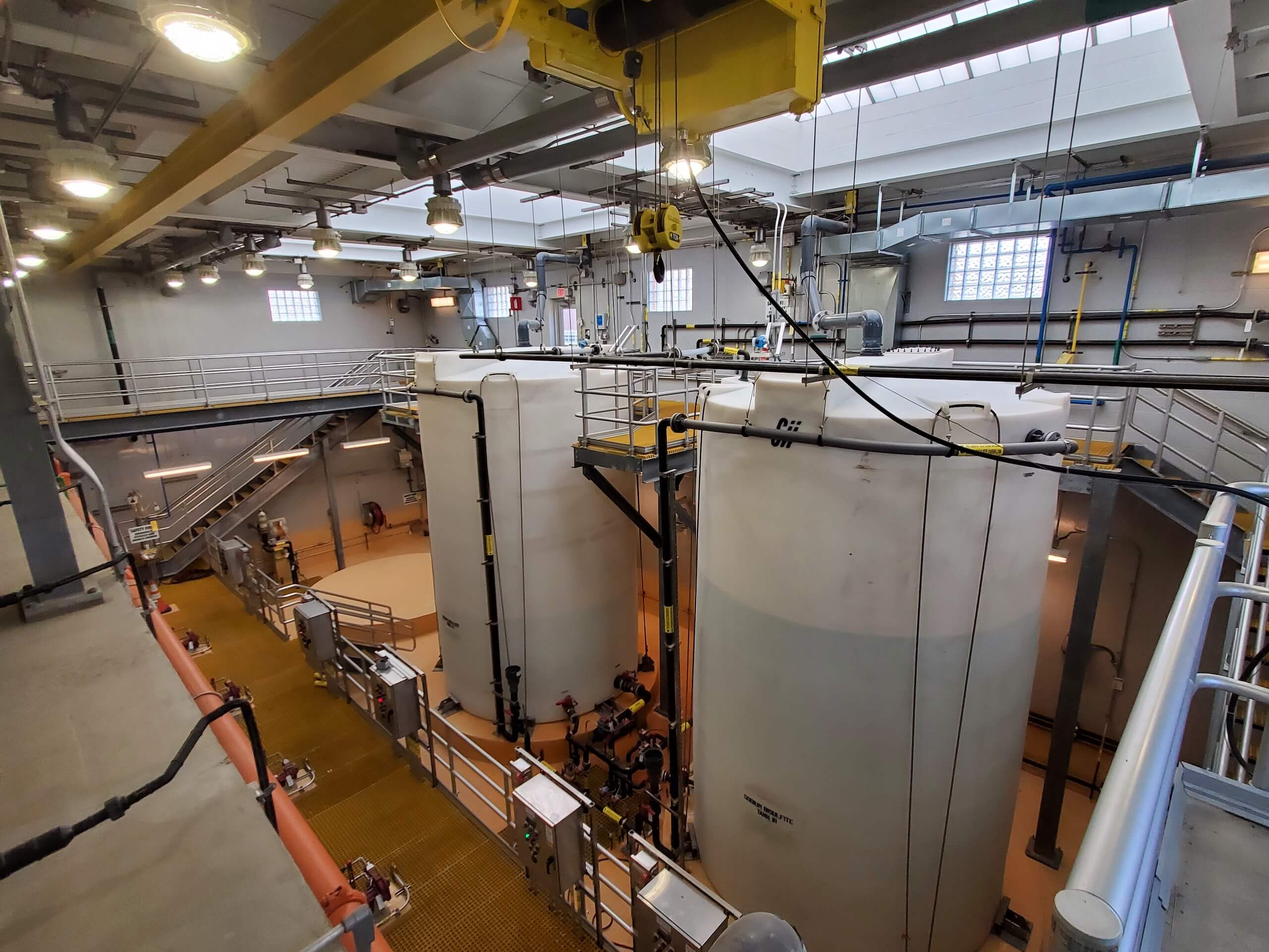 A large industrial facility interior with three tall, white storage tanks, overhead cranes, metal walkways, pipes, and industrial lighting, viewed from an elevated platform.