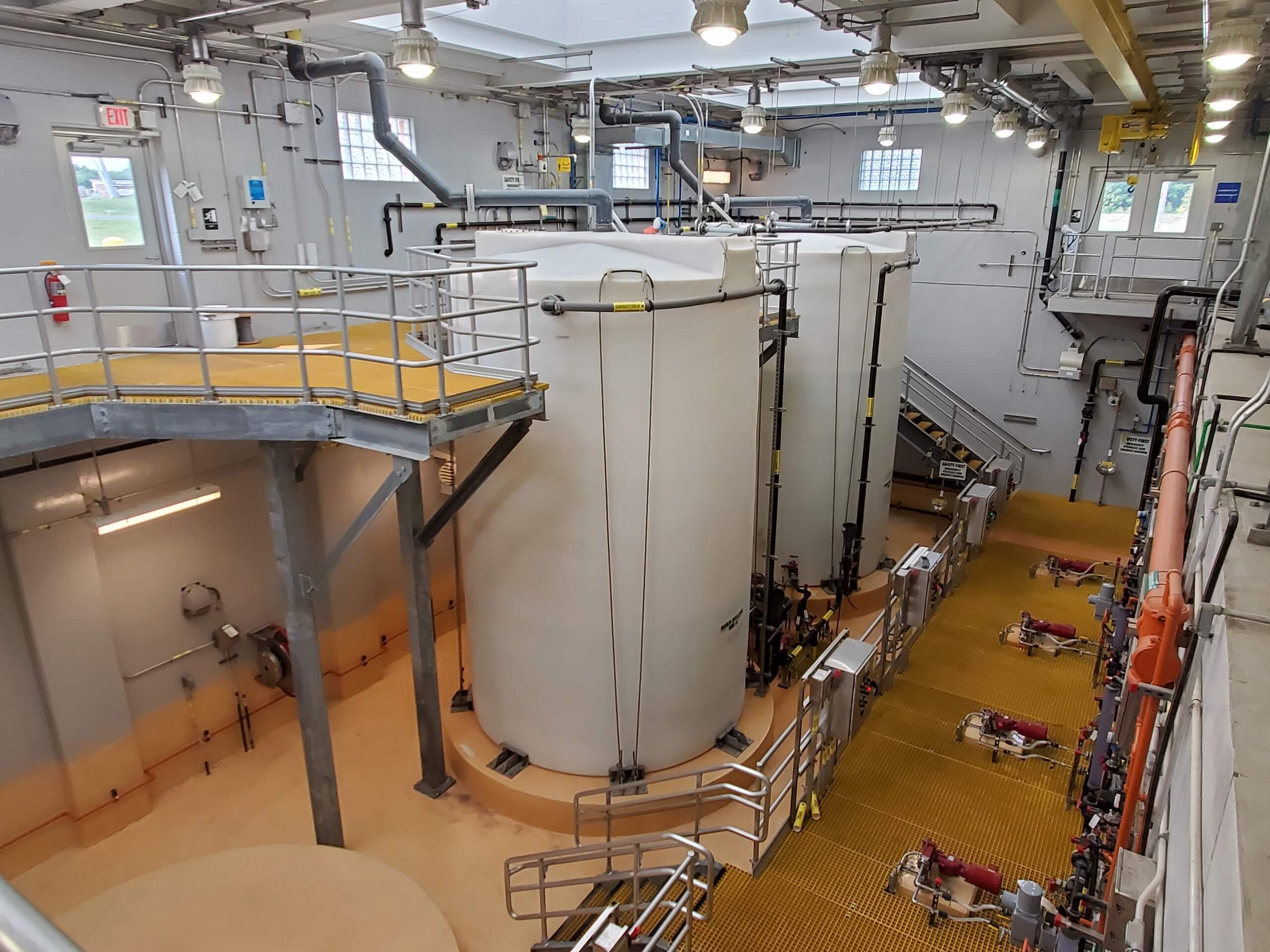 Large white industrial tanks inside a facility with metal railings, overhead lights, and yellow grated walkways. Pipes and machinery are visible on the sides and floor, with stairs leading to an upper level.