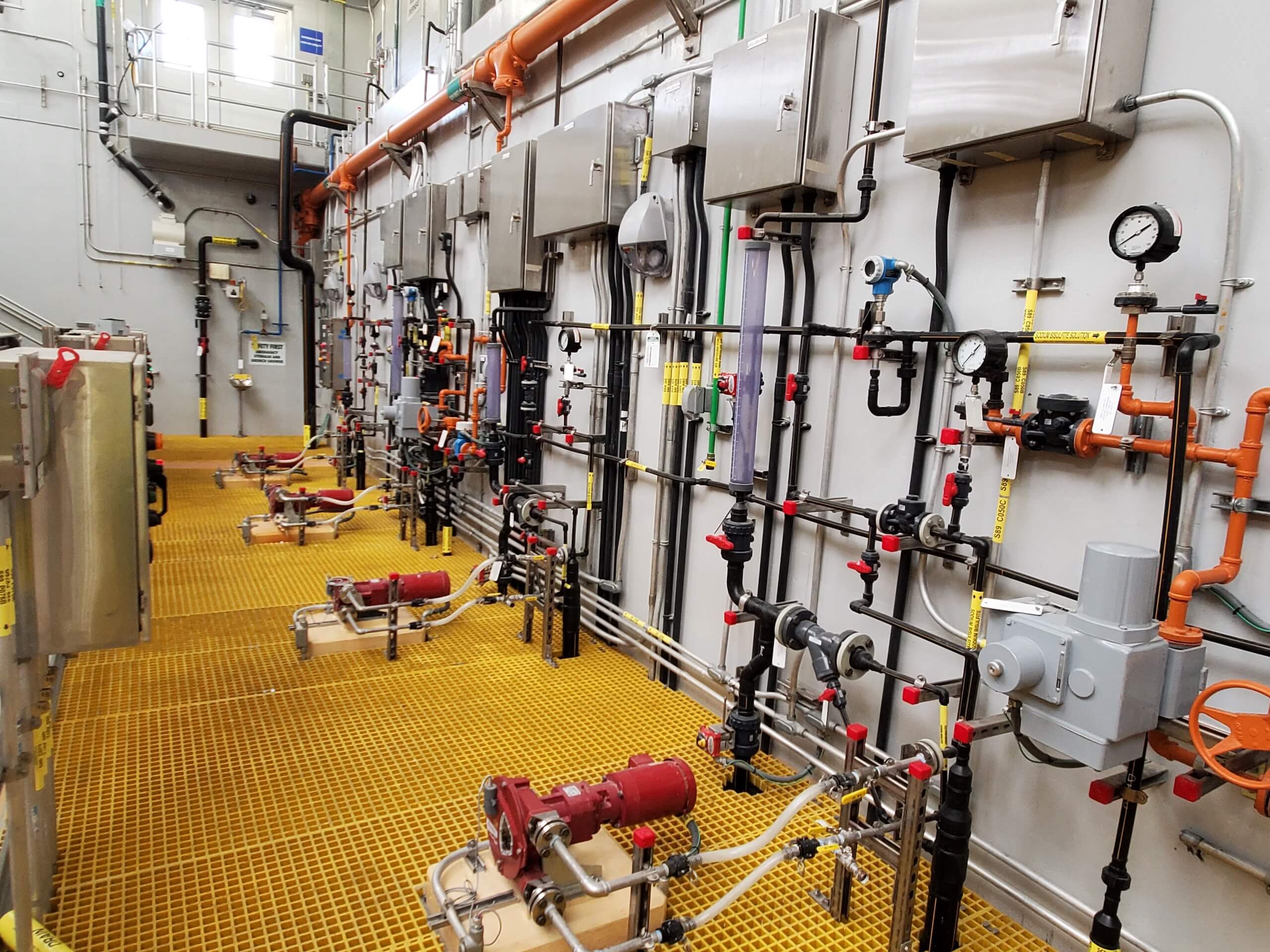 A mechanical room with various pipes, gauges, valves, and control panels mounted on the wall, above a yellow grated floor. The pipes include orange, black, and silver sections.
