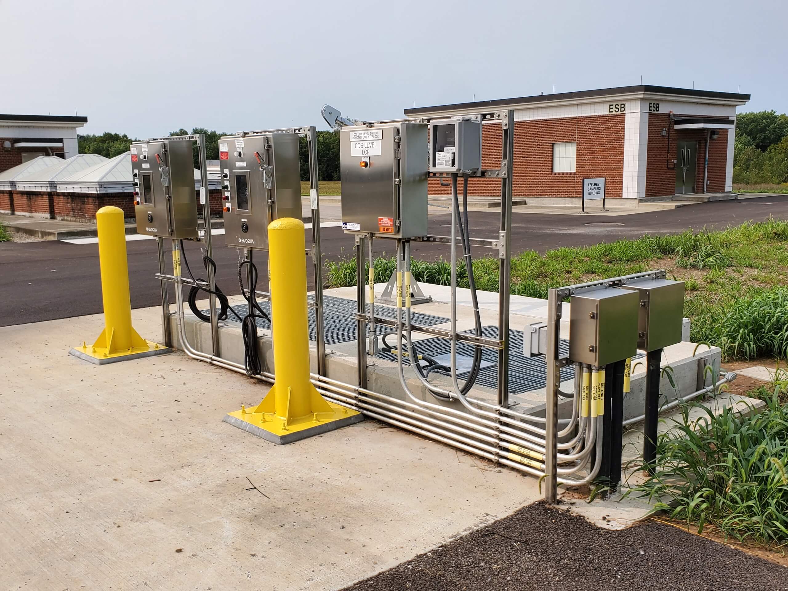 Outdoor utility station with metal control panels, conduits, and yellow protective bollards on a concrete pad. Two brick buildings are visible in the background, with grass and pavement surrounding the area.