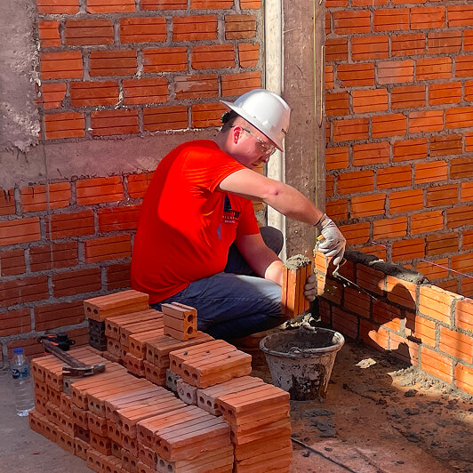 A construction worker wearing a white hard hat and red shirt is building a brick wall, placing bricks with mortar. Stacks of bricks and a bucket are nearby.