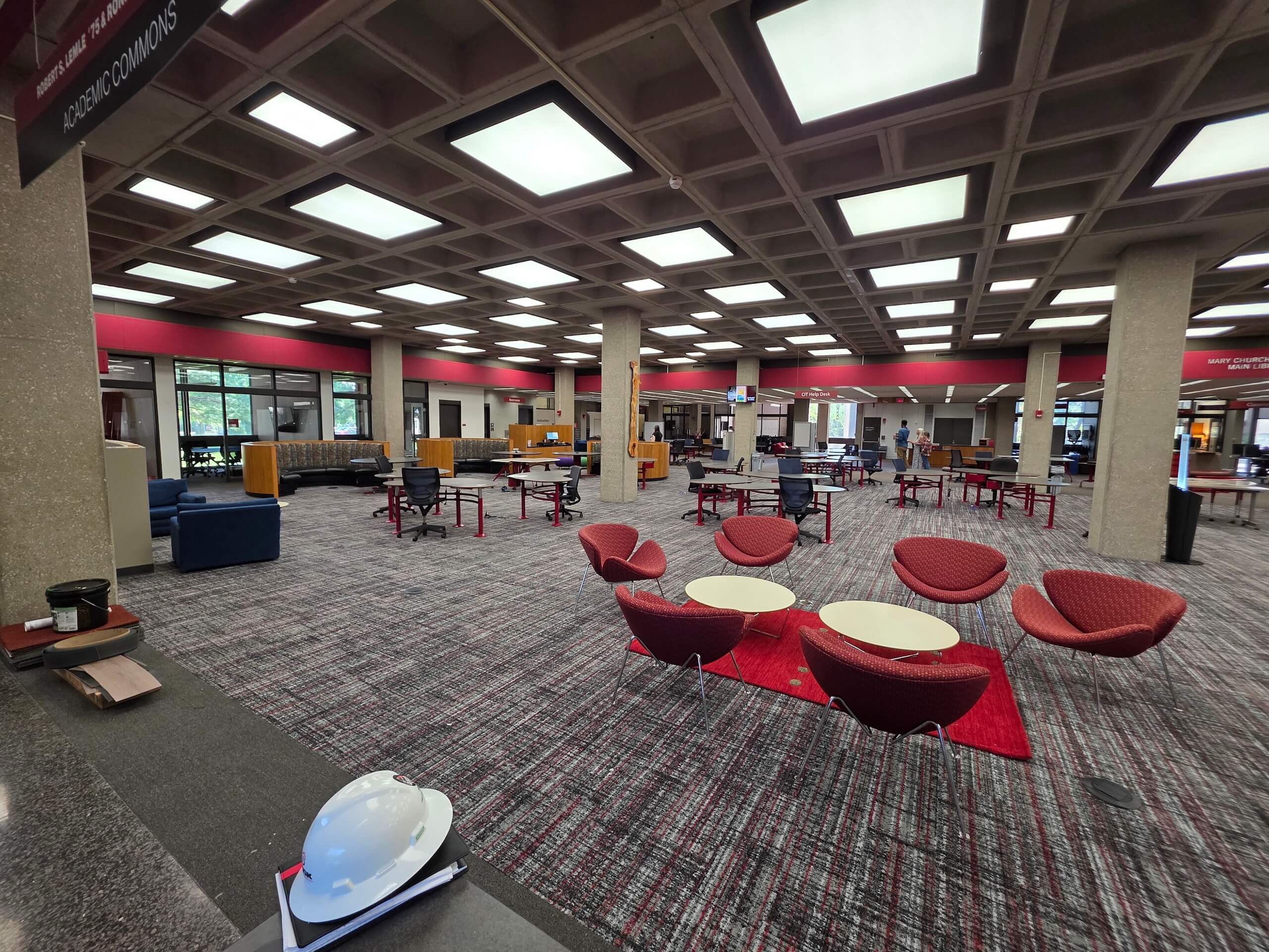 A spacious library commons area with red chairs, round tables, carpeted floor, and large ceiling lights. People are scattered throughout, working or walking. Large windows provide natural light.
