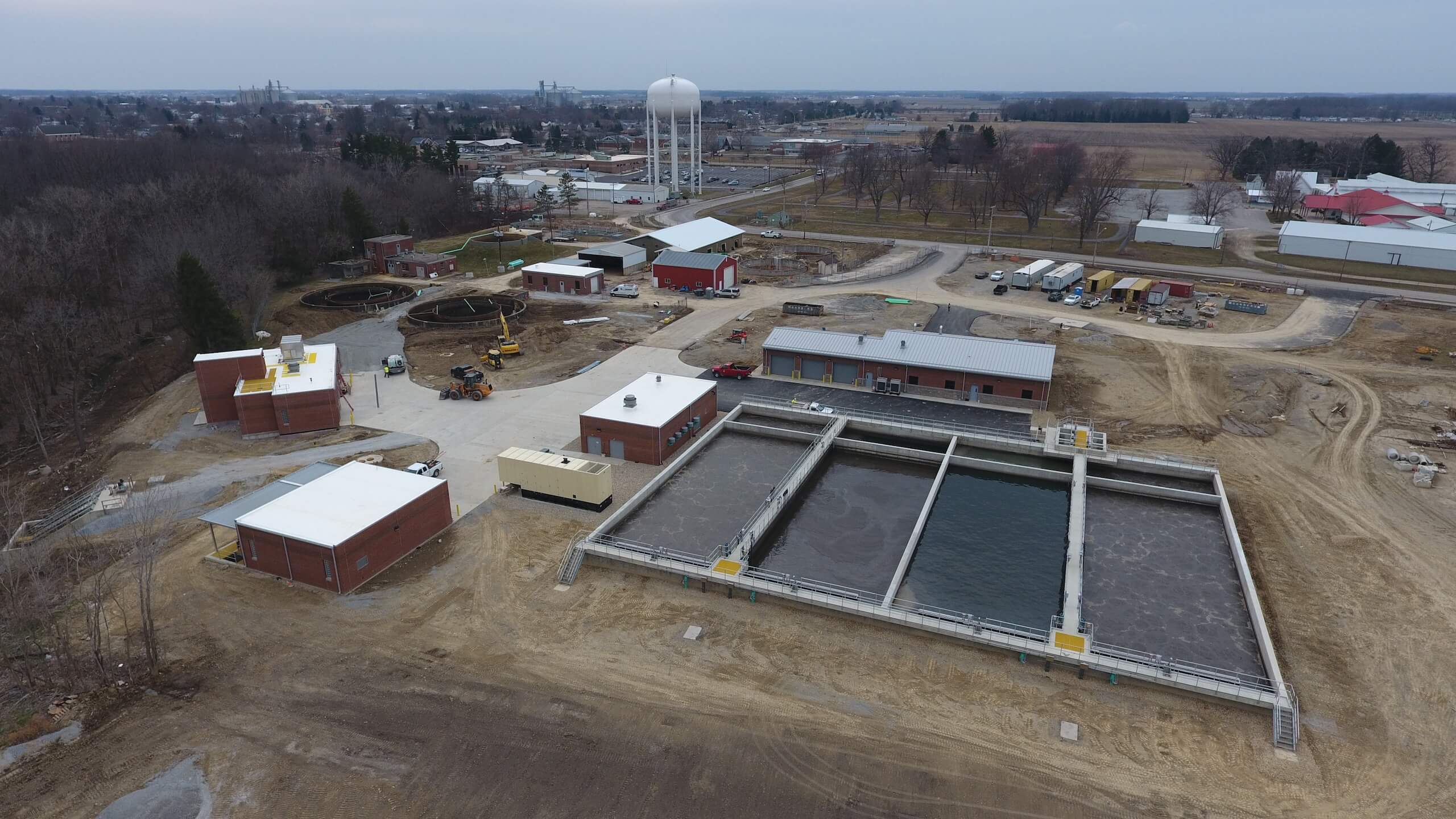 Aerial view of a water treatment facility with large tanks, rectangular settling basins, multiple brick buildings, and a water tower in the background, surrounded by a rural landscape and scattered trees.