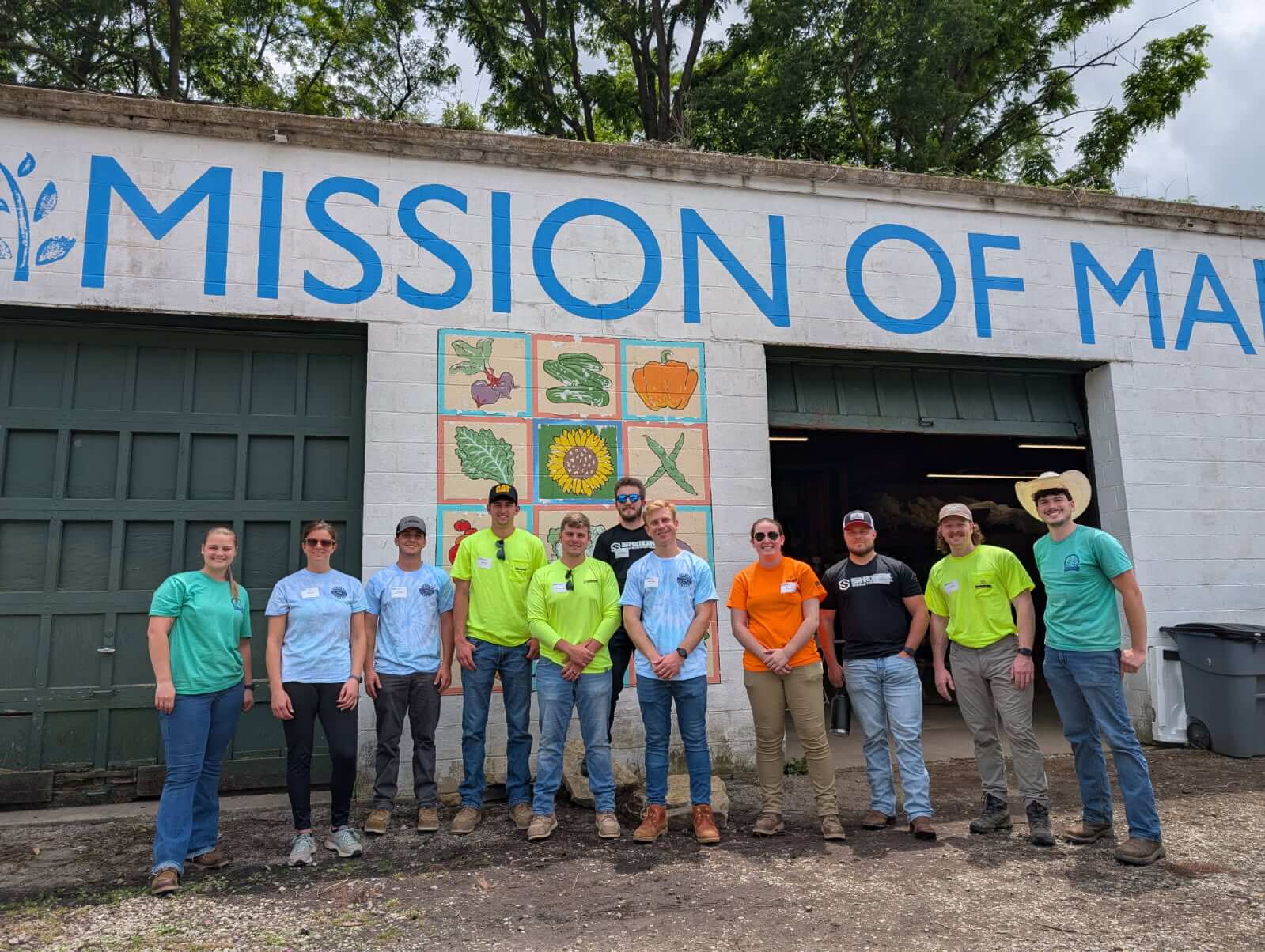 Twelve people stand in front of a building with MISSION OF MA painted on it and colorful produce art. They wear casual work clothes and pose together, smiling outdoors.