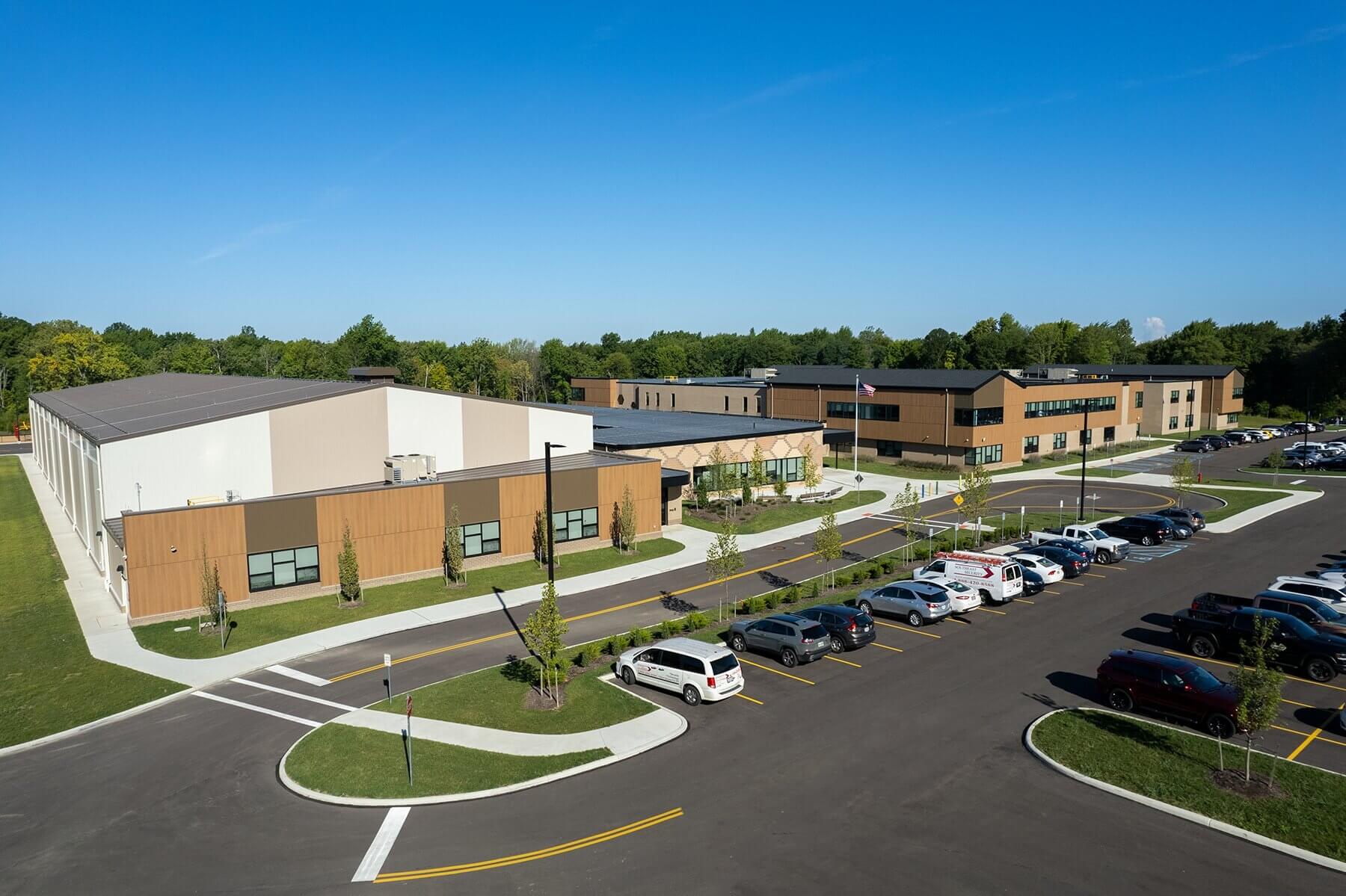 A modern school building with large windows and tan walls is surrounded by trees. Several cars are parked in the adjacent lot under a clear blue sky.