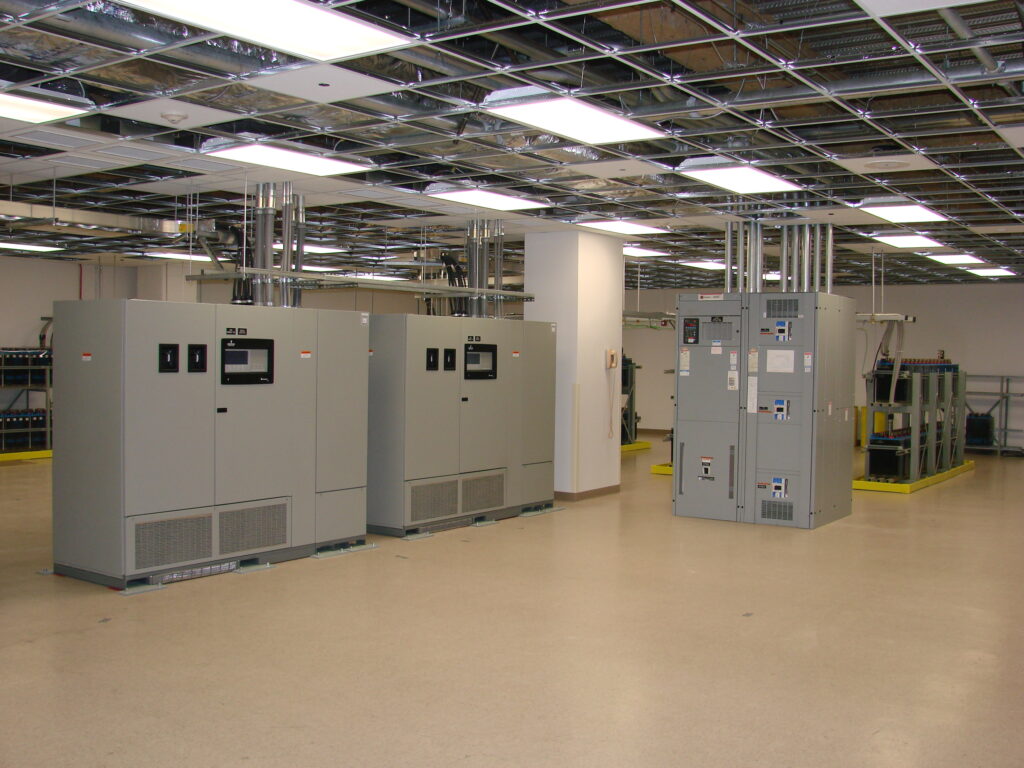 A large, clean room with electrical control panels, open ceiling showing wiring, and various industrial equipment along the walls. The floor is beige and shiny.