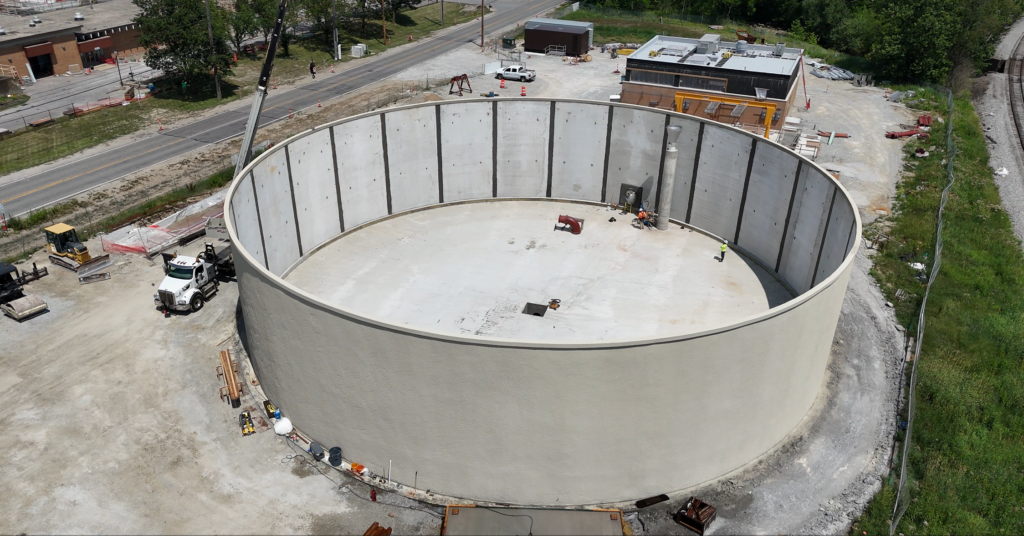 Aerial view of a large, circular concrete structure under construction, with workers and equipment inside. Nearby are buildings, a road, and some grassy areas.