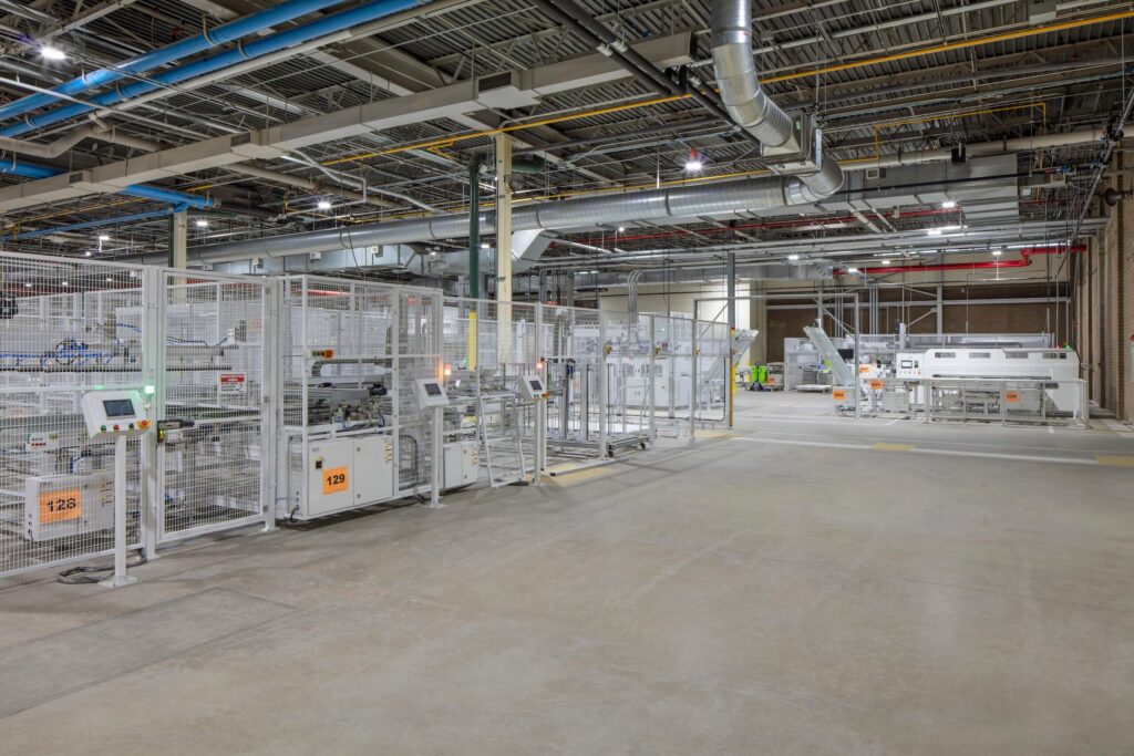 A spacious, modern factory floor with automated machinery, white safety barriers, exposed pipes, and ductwork overhead. The area is clean, well-lit, and mostly empty, with industrial equipment lining the sides.