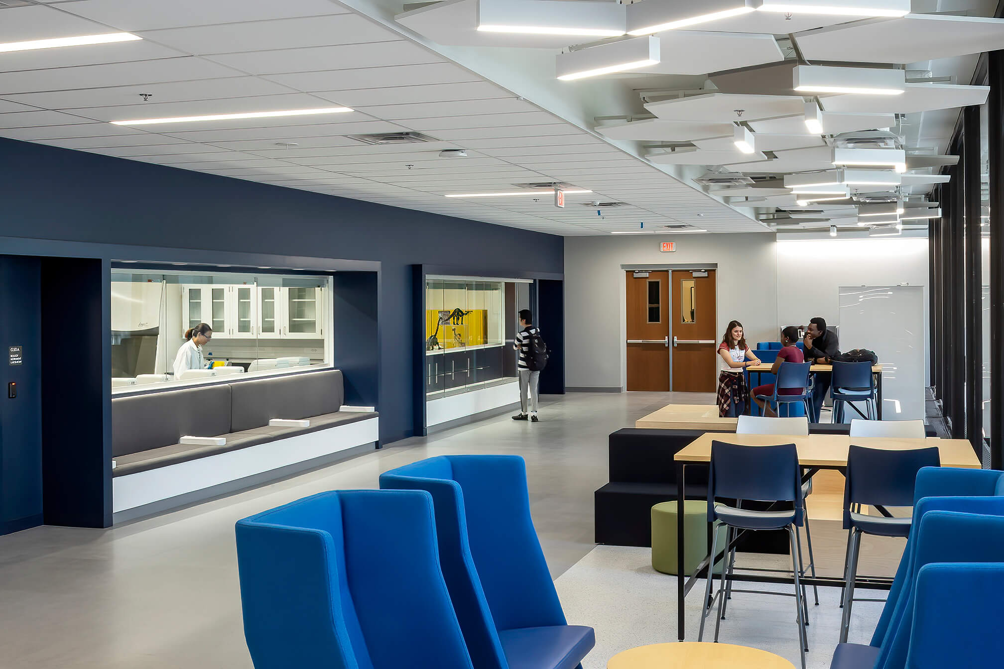 Students sit and stand in a modern, open common area with blue chairs and tables, near large windows and a counter where a staff member works behind glass. Ceiling panels are geometric, and lighting is bright.
