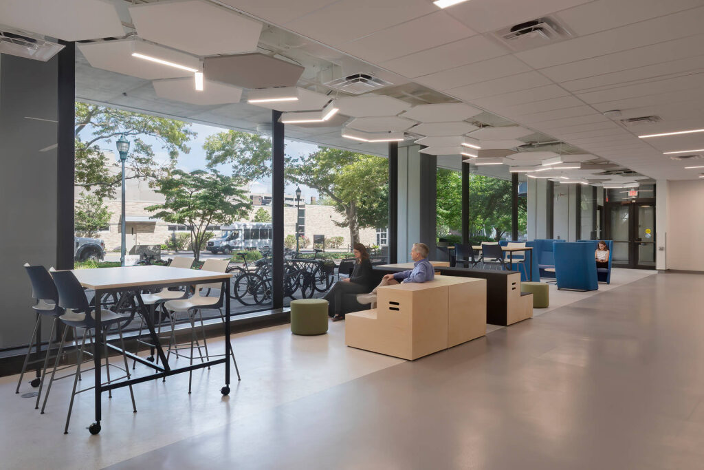 Two people sit and talk in a modern, open indoor space with large windows, high tables, and natural light, overlooking trees and a row of bicycles outside.