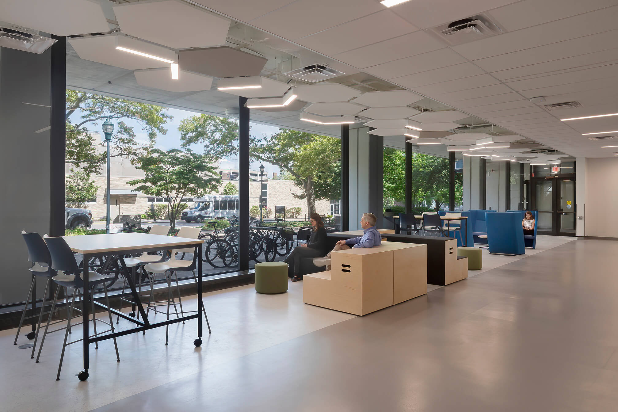 Two people sit and talk in a modern, open indoor space with large windows, high tables, and natural light, overlooking trees and a row of bicycles outside.