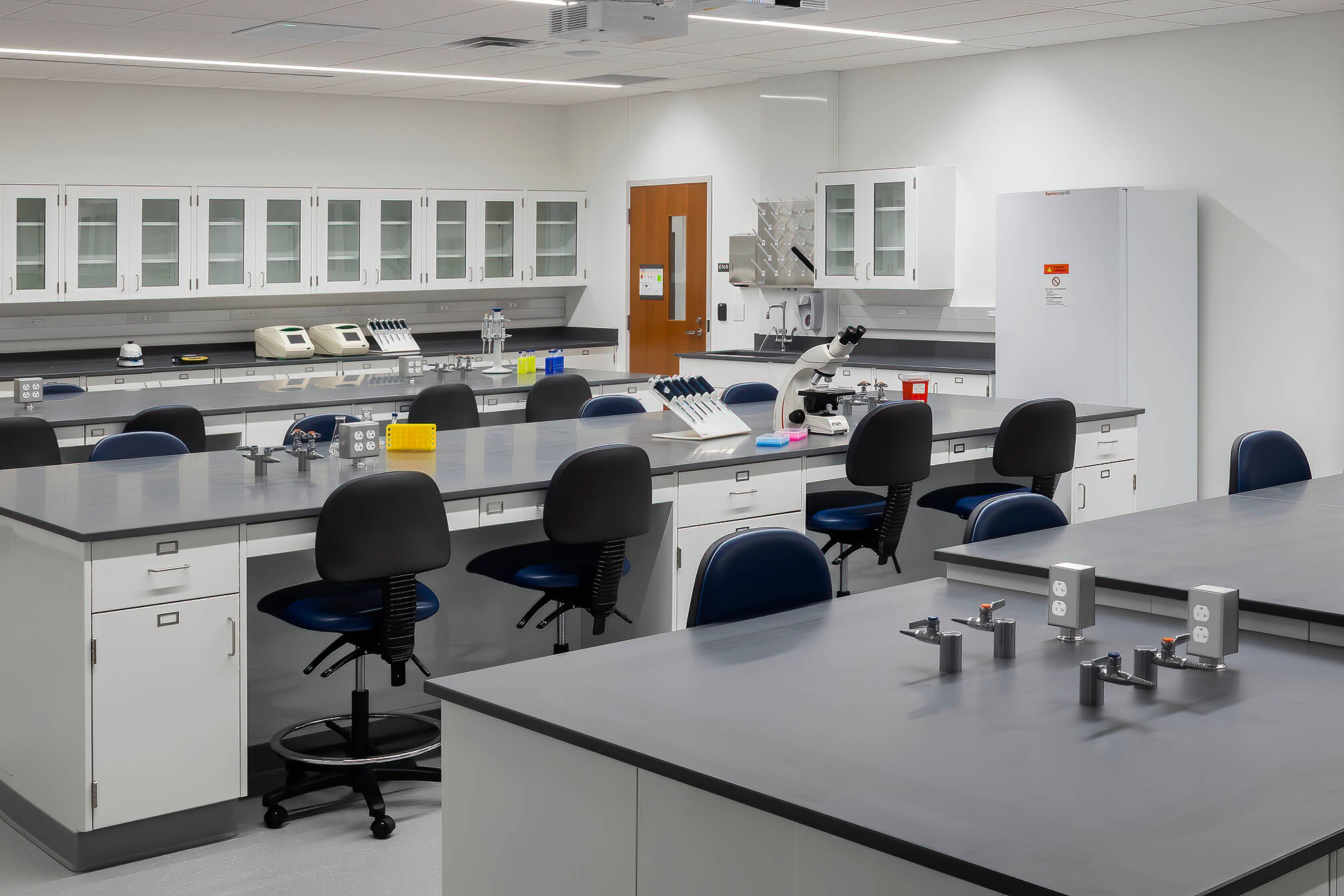 Modern science laboratory with workstations, lab chairs, microscopes, equipment on counters, cabinets, and a refrigerator. The space is clean, organized, and well-lit with white walls and grey countertops.