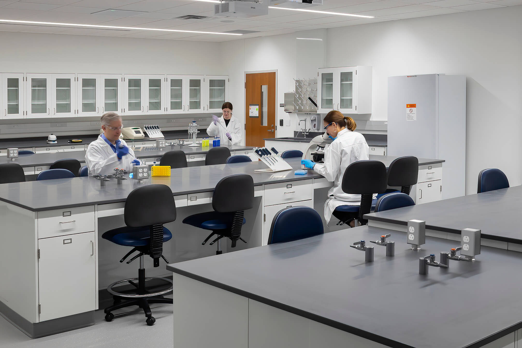 Three people in lab coats work at laboratory benches with scientific equipment and computers. The lab is clean, with white walls, glass cabinets, and a refrigerator in the background.