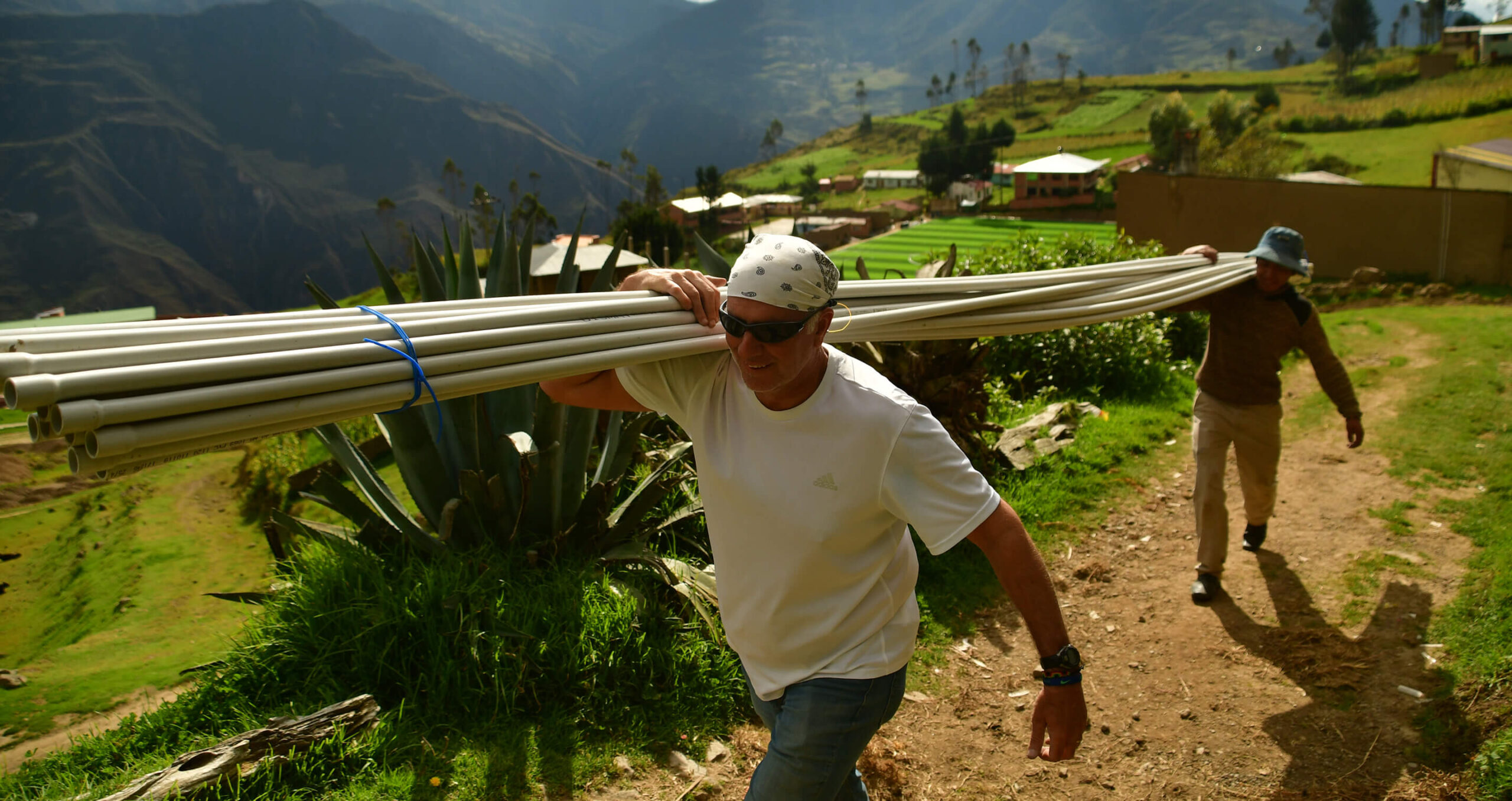 A man wearing sunglasses and a bandana carries long pipes on his shoulder along a dirt path in a rural, mountainous area. Another person walks behind him, and green fields and scattered buildings are visible in the background.