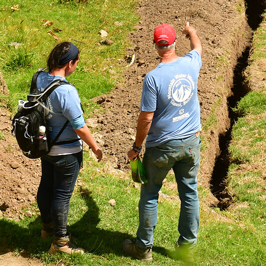 Two people in blue shirts stand next to a freshly dug trench outside. One points ahead while holding papers; the other listens, carrying a backpack and water bottles. Grass and dirt are visible around the trench.
